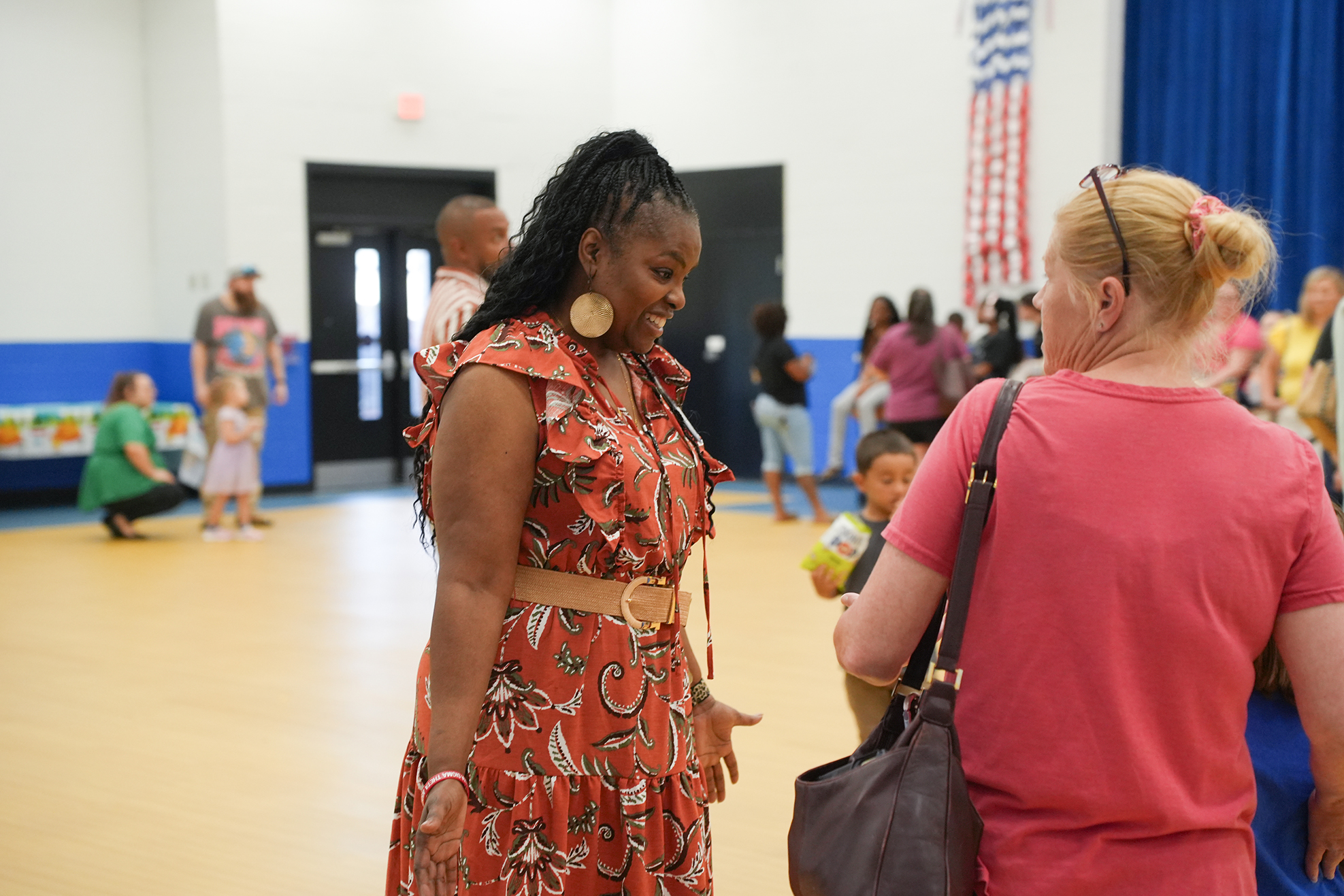 A woman in a patterned dress speaks with another woman wearing a pink top in an indoor space.