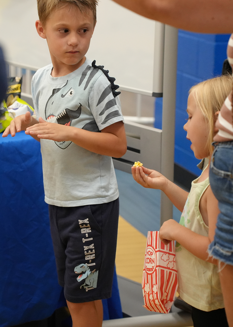 Two children face each other in a gymnasium; one holds popcorn and the other a dinosaur-print T-shirt.