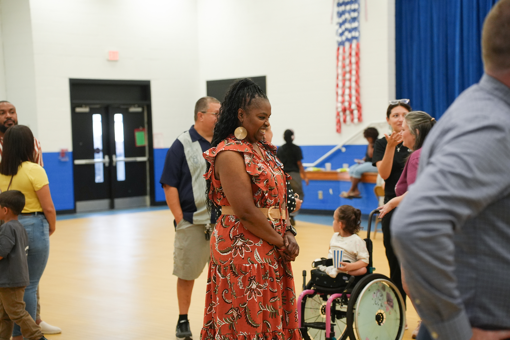 A woman in a patterned dress is standing with a group of people in a hall. A child in a wheelchair is nearby.