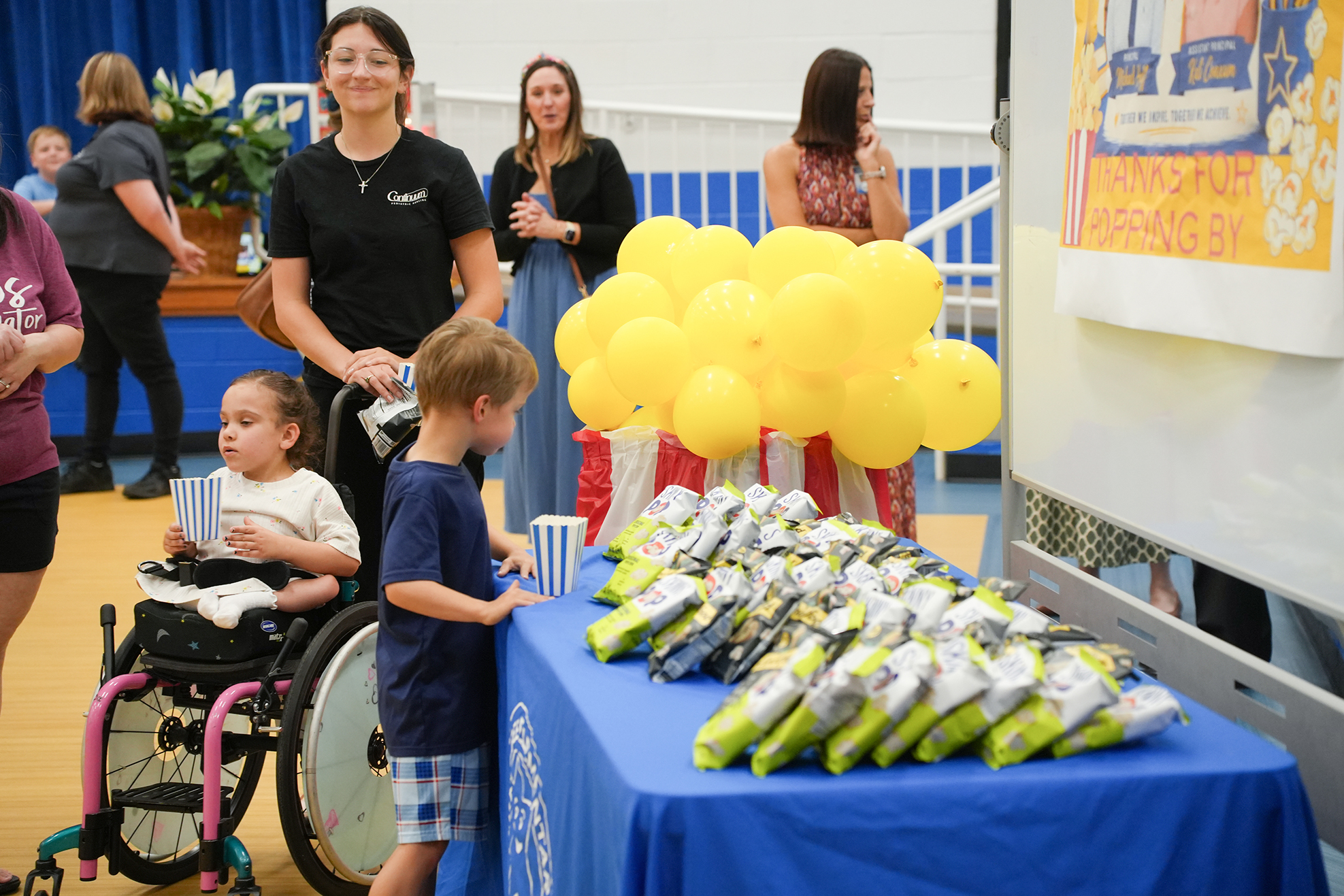 A table with blue cover displays yellow balloons and food. People are standing around it, including a woman in a wheelchair.