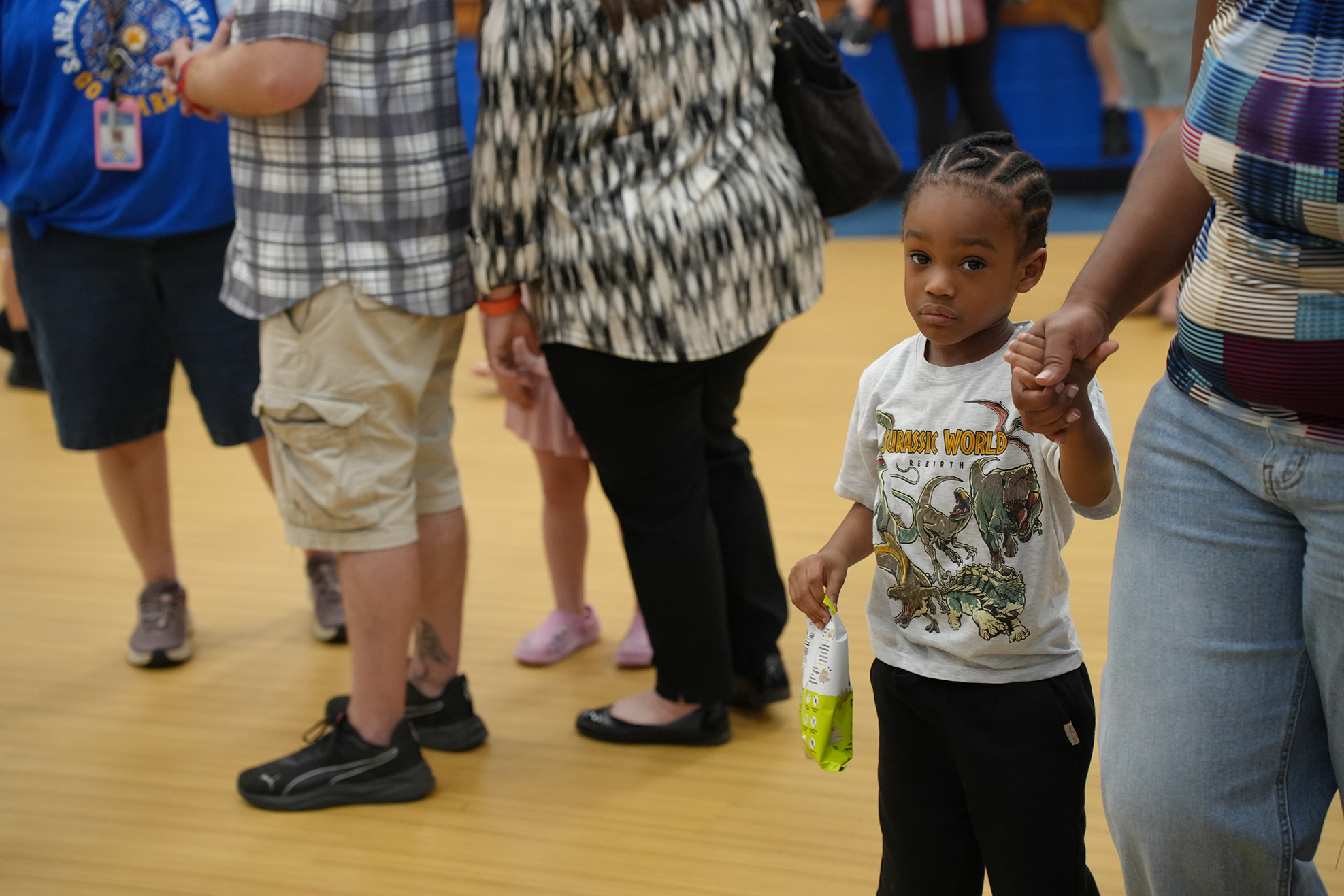 A small child with a dinosaur t-shirt holds hands with an adult, while others walk on a wooden floor.