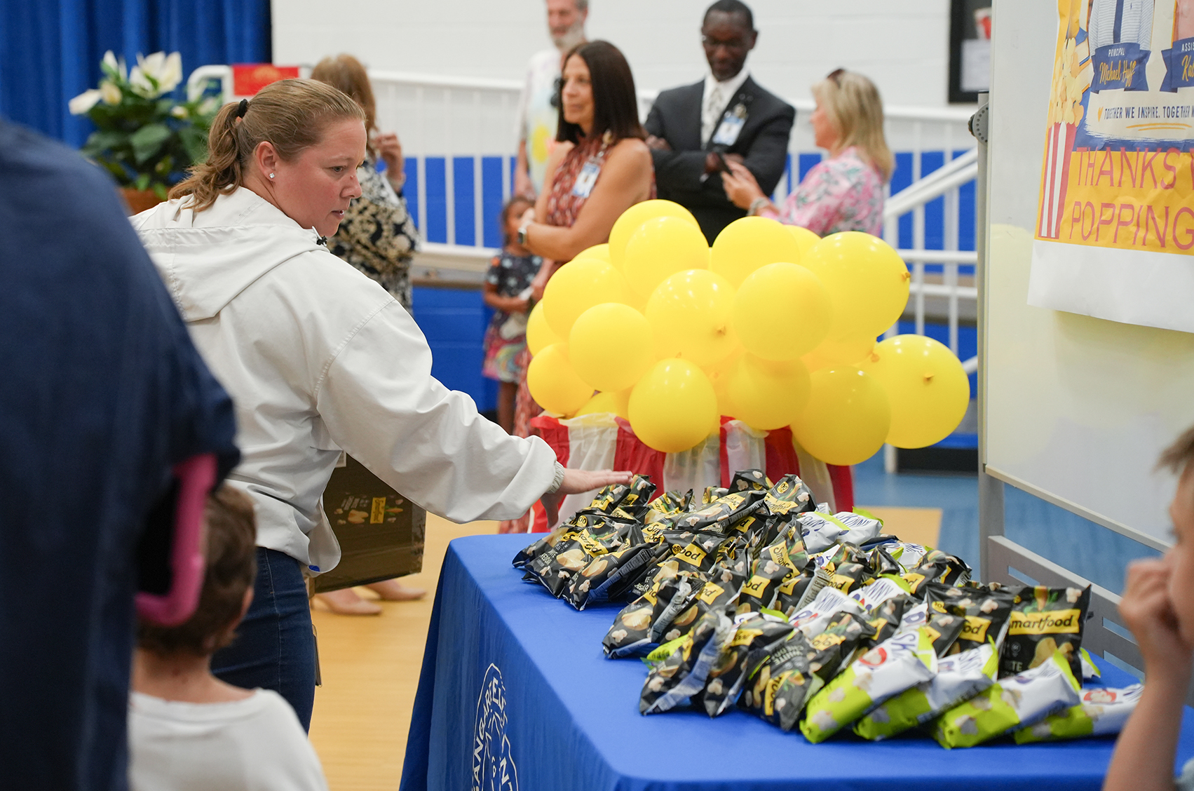 People gather around a table with a yellow balloon display and bags. One woman hands out items from the table.