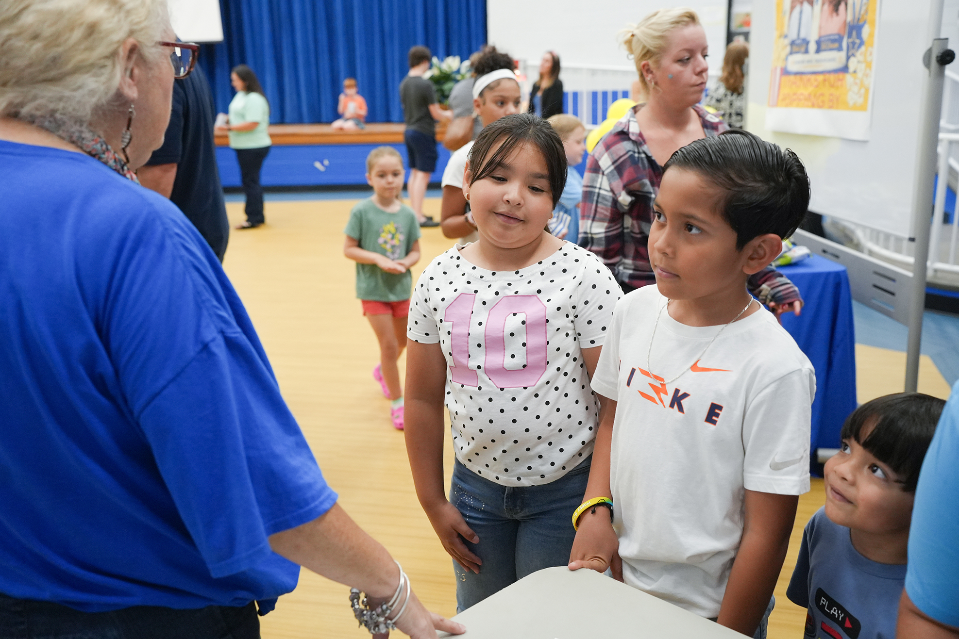 Several children stand in a gymnasium with a woman, one with a polka-dot shirt, another in a Nike shirt.