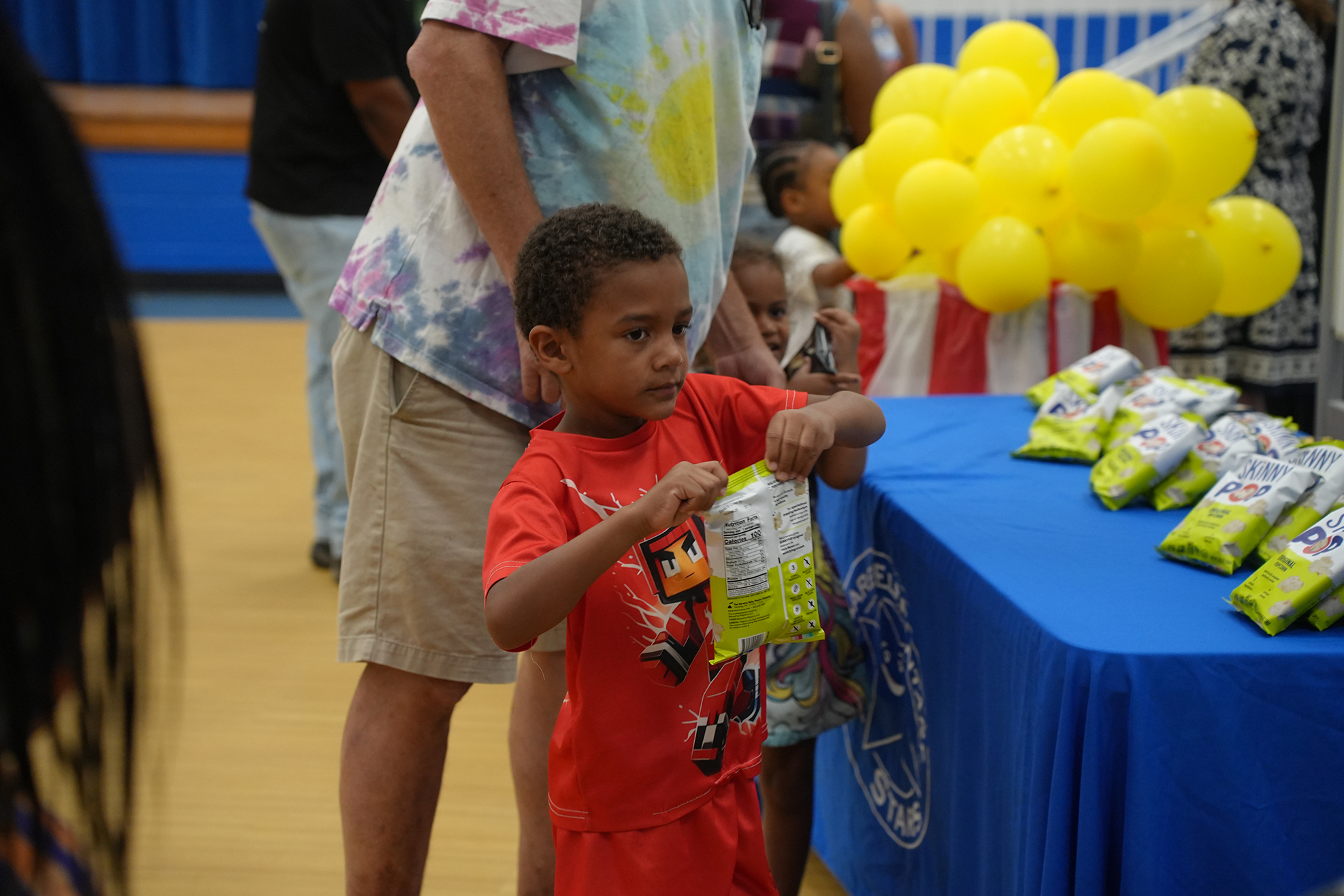 A young boy in red holds a bag of food, standing next to an adult at a table with balloons and snacks.