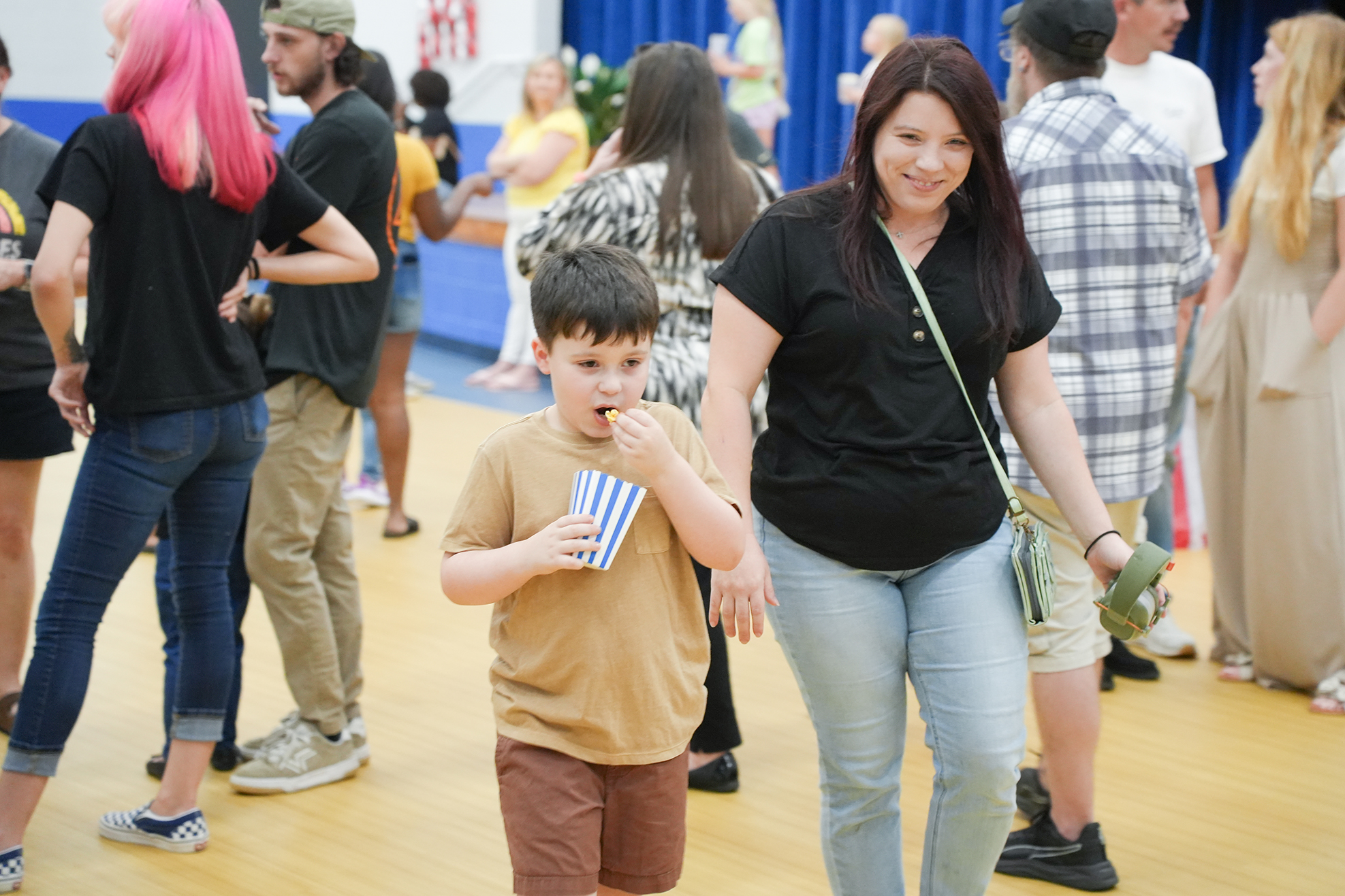 A woman and boy walk through a hall filled with people. The boy eats a treat and holds a paper cup.