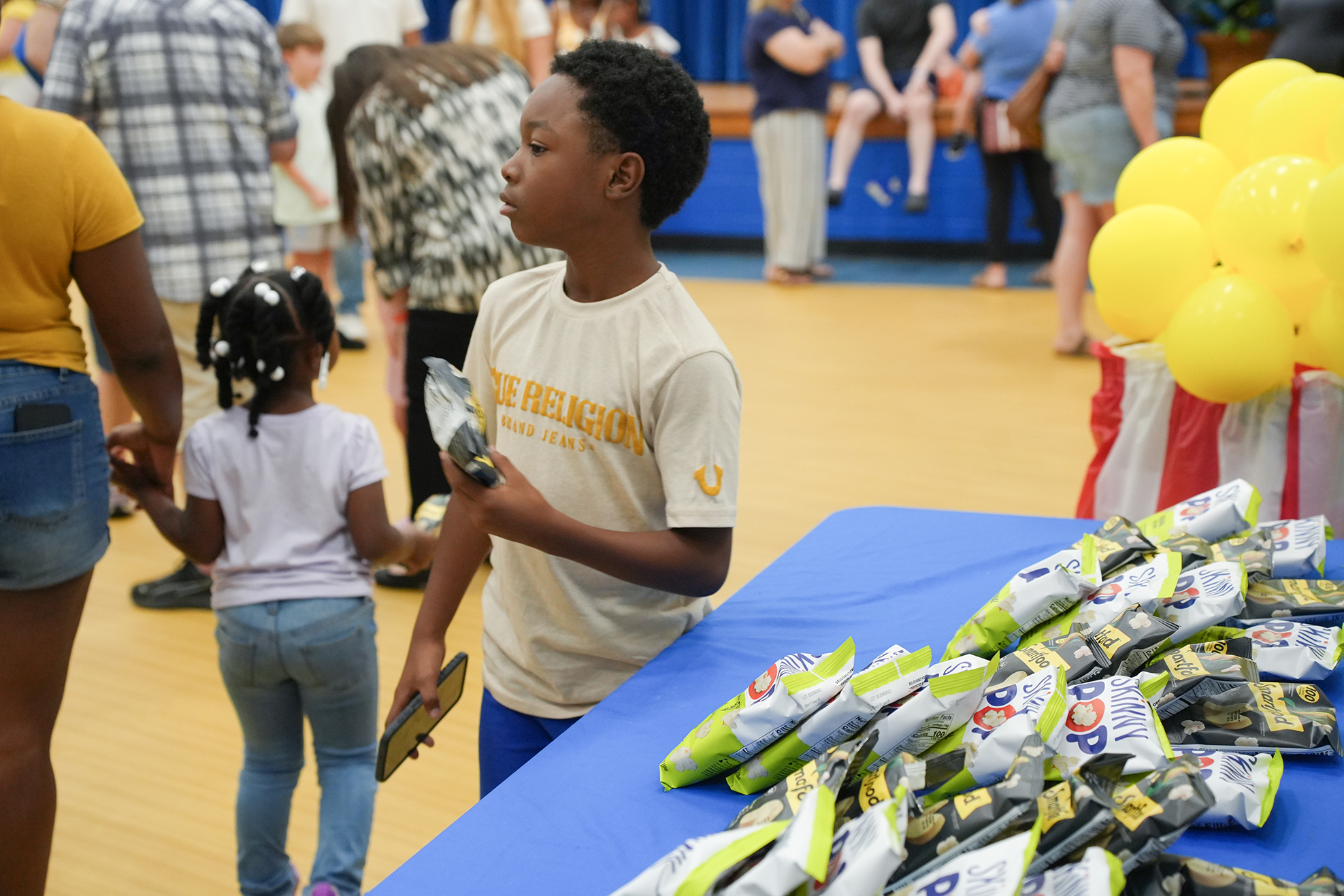 A young boy holds an award while a girl holds his hand. A table has bags of food.