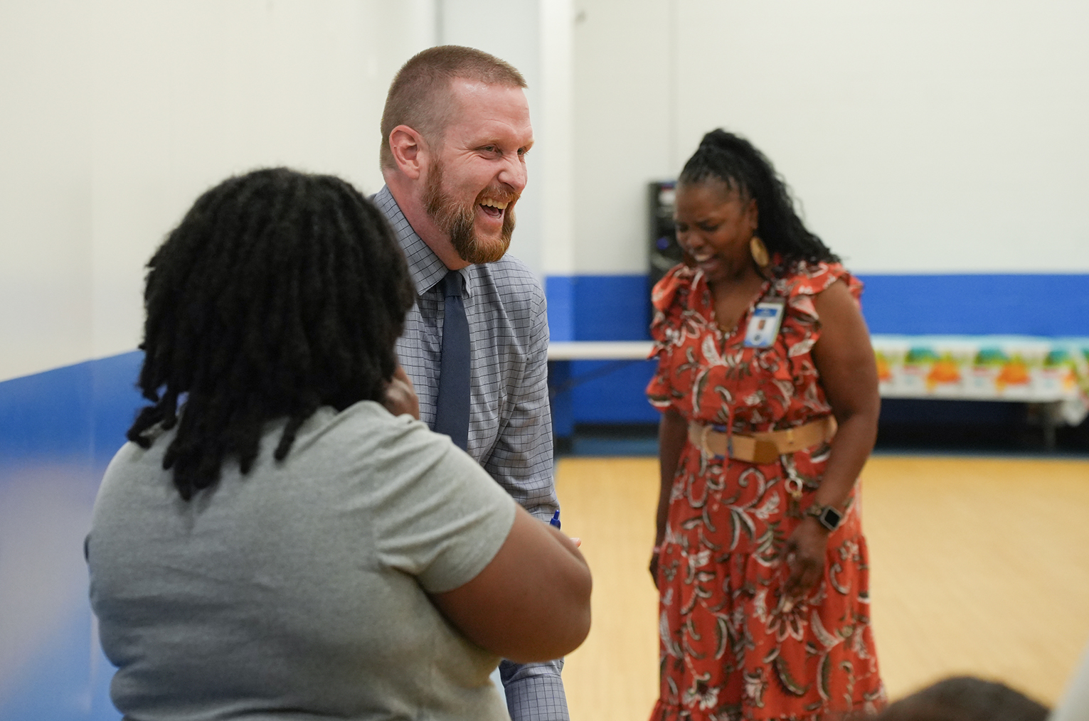 Three people in a room with blue and white walls and a wooden floor. A woman in a dress and a man in a tie laugh.