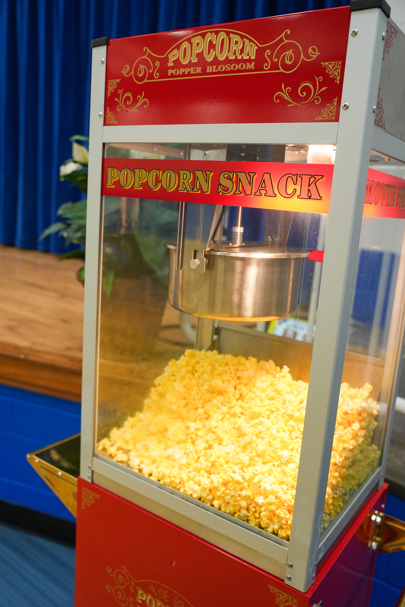 A popcorn machine with a red and silver exterior, clear glass window, and red label reading "Popcorn Snack." Popcorn inside the machine. Background includes a blue curtain and wooden surface.