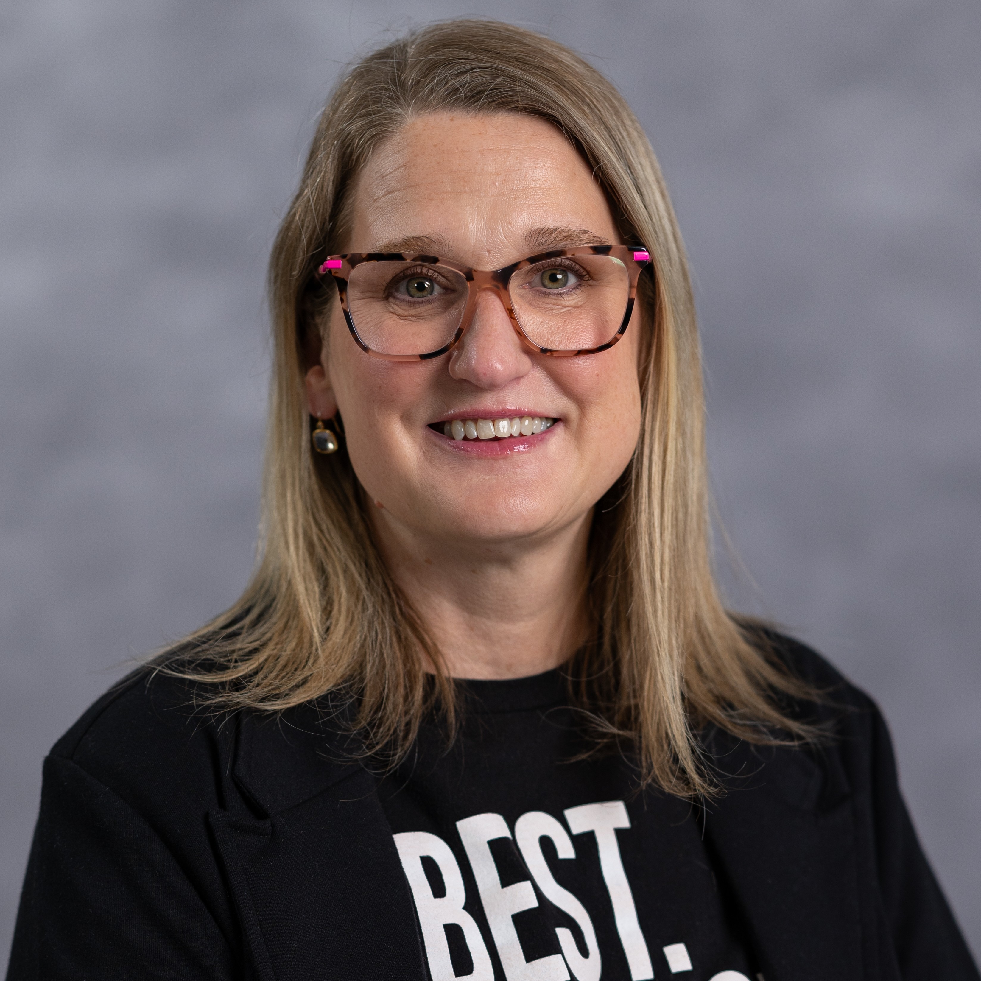 A woman with blonde hair and glasses smiles. She wears a black shirt with white text that reads "BEST. SCHOOL."