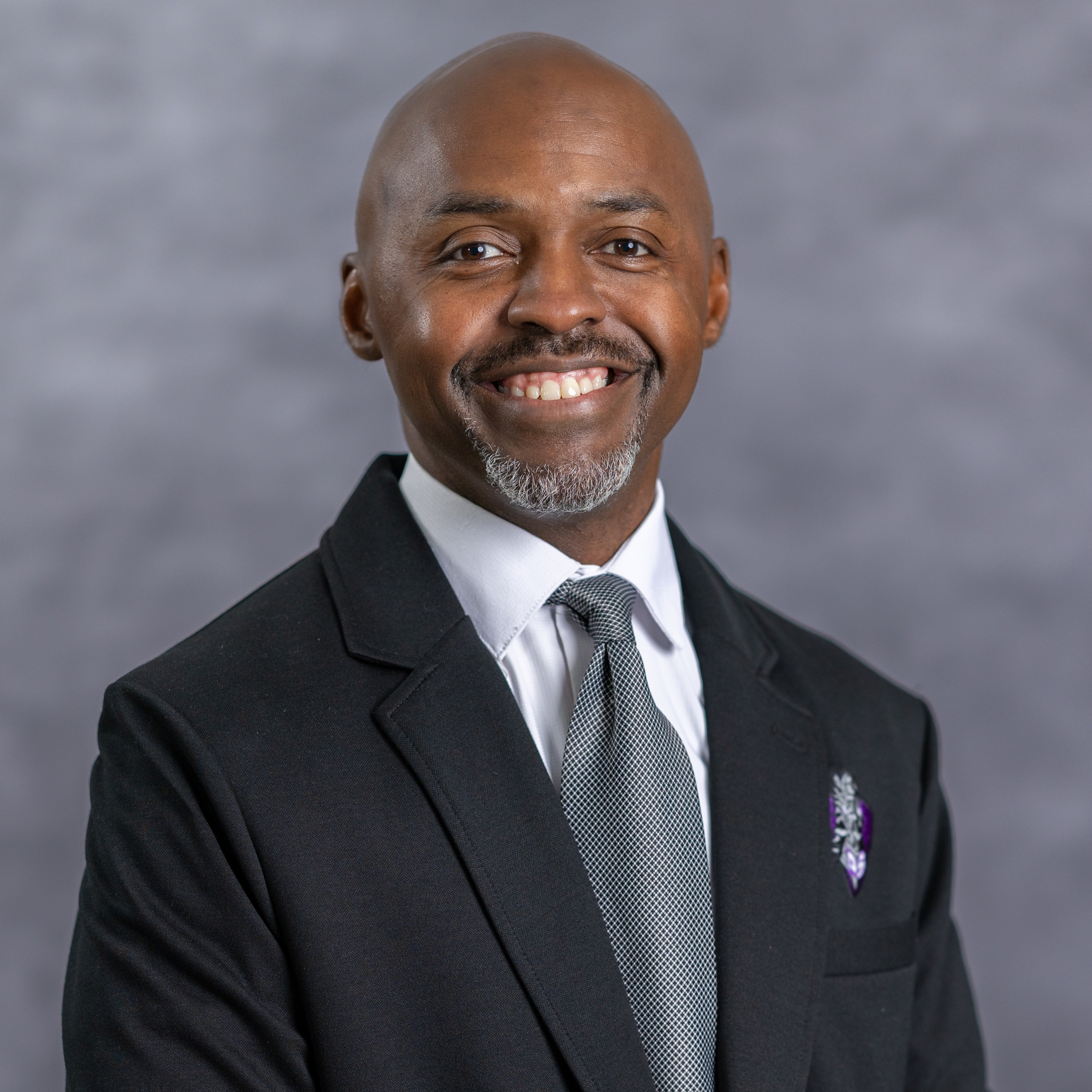 A man with a smile and gray tie pin stands against a gray background, wearing a dark suit.