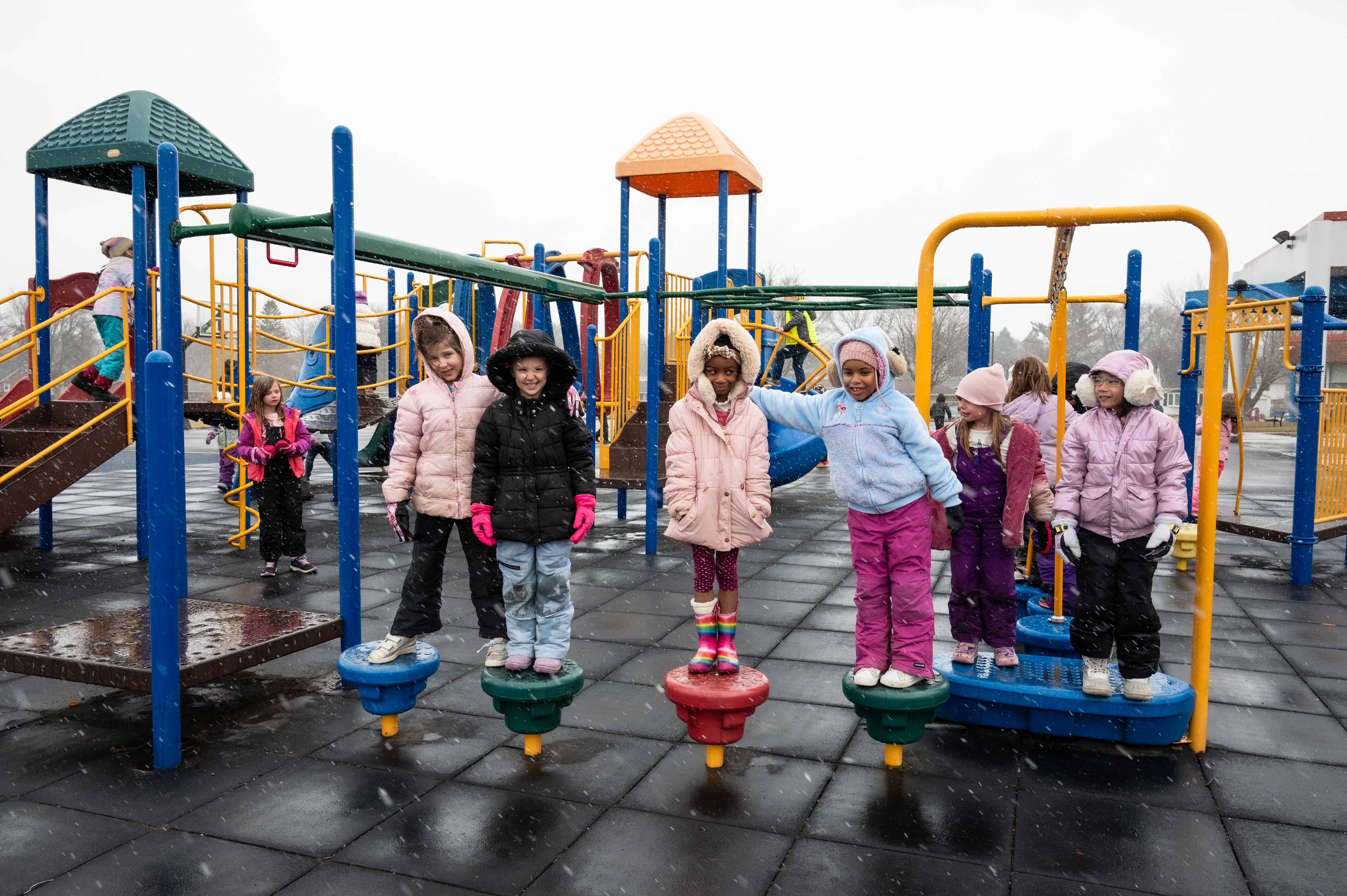 Children bundled up in winter gear stand on the playground equipment and smile at the camera.