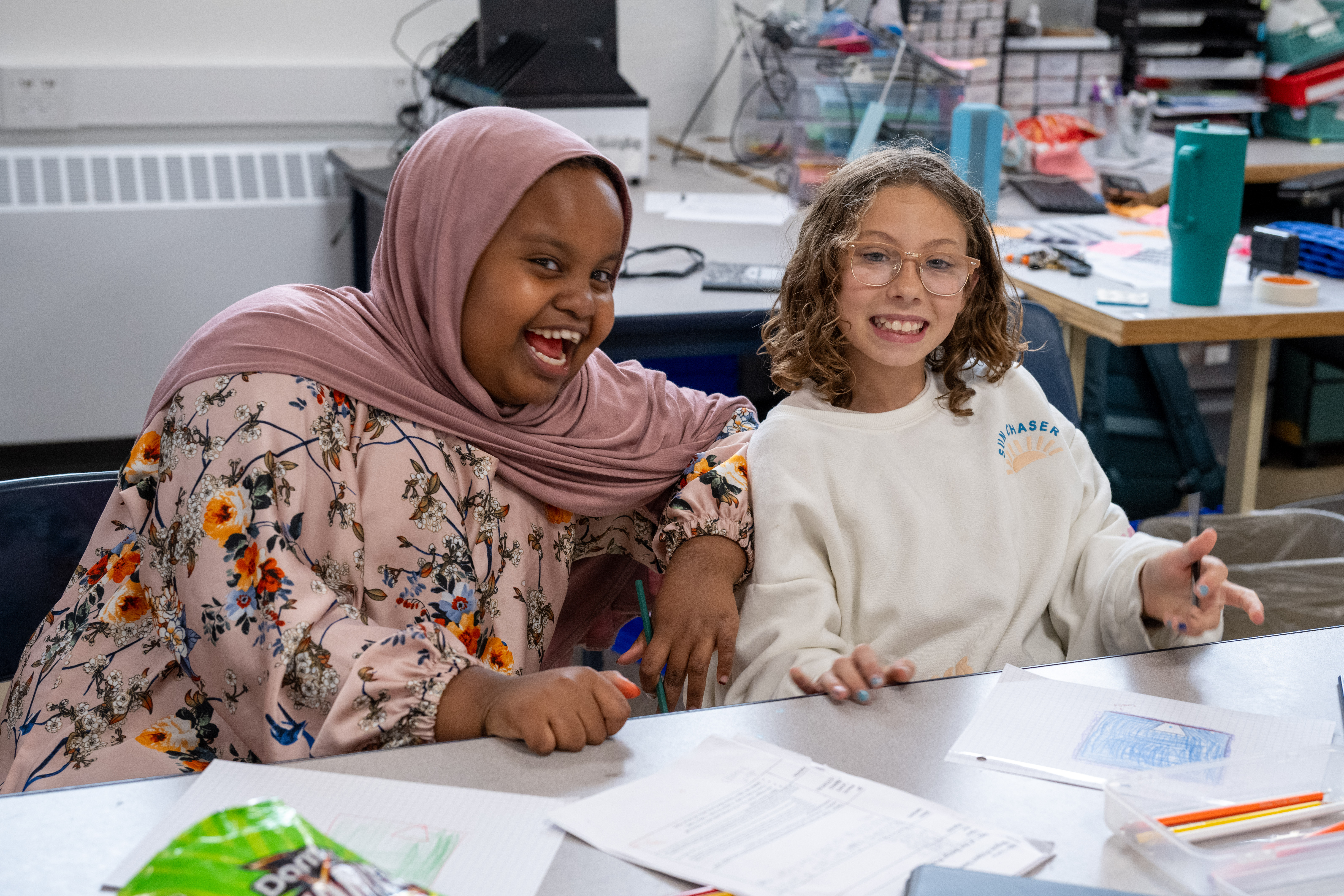 Two smiling students sit side by side at a classroom table with papers and colored pencils, laughing while working on an activity together.