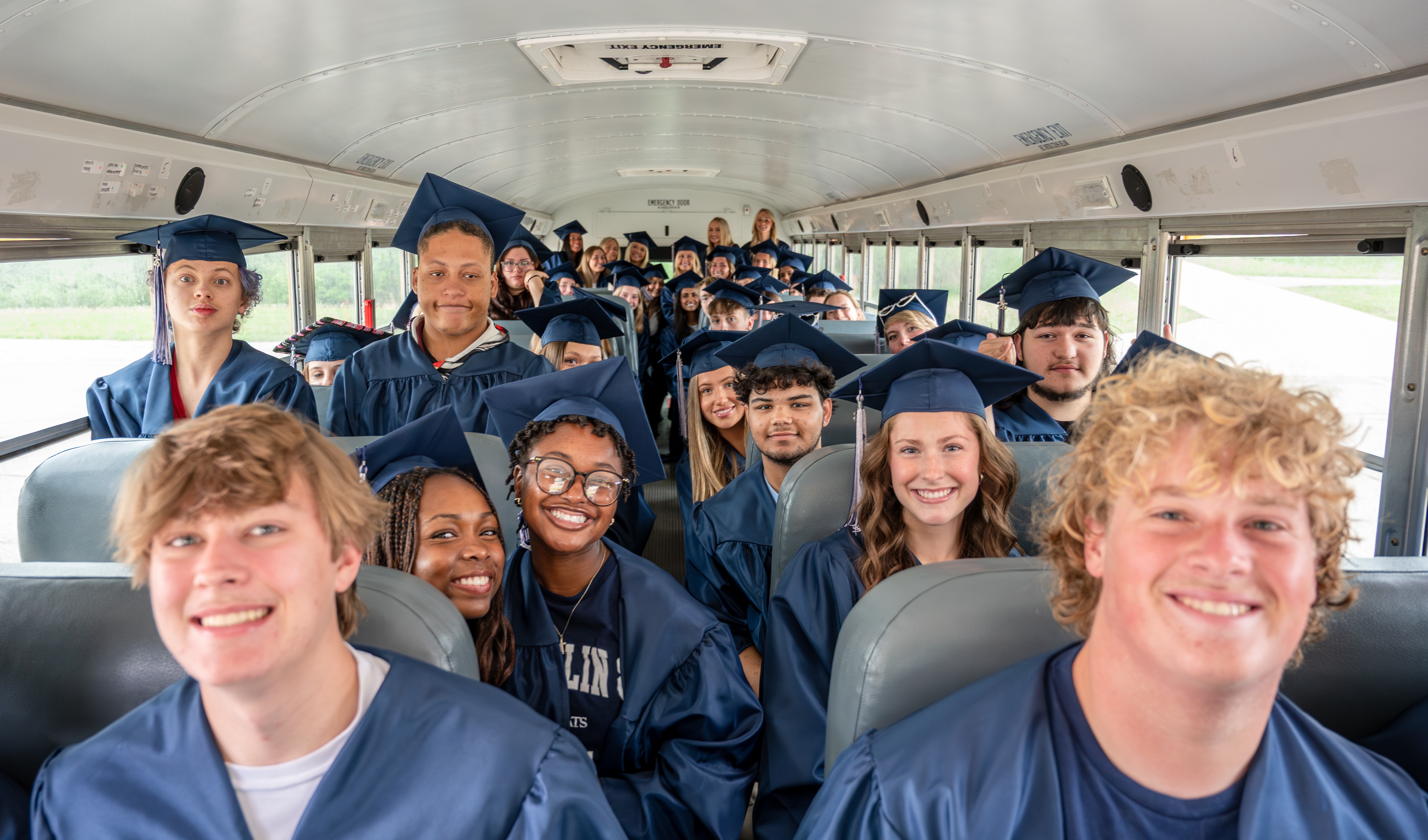 Seniors in caps and gowns inside a school bus look towards the front of the bus and smile for camera.
