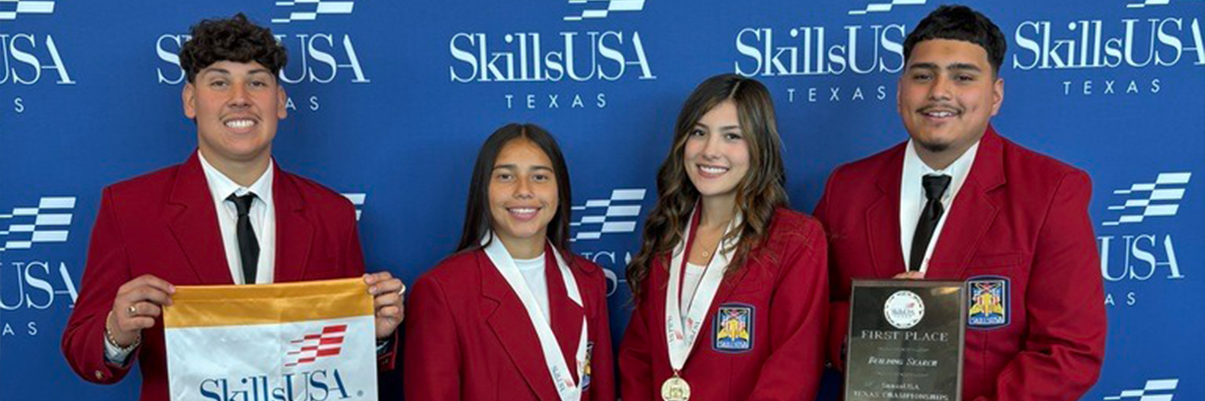 Four people in red coats and ties, possibly students, stand together, holding a flag, medals, and a plaque.