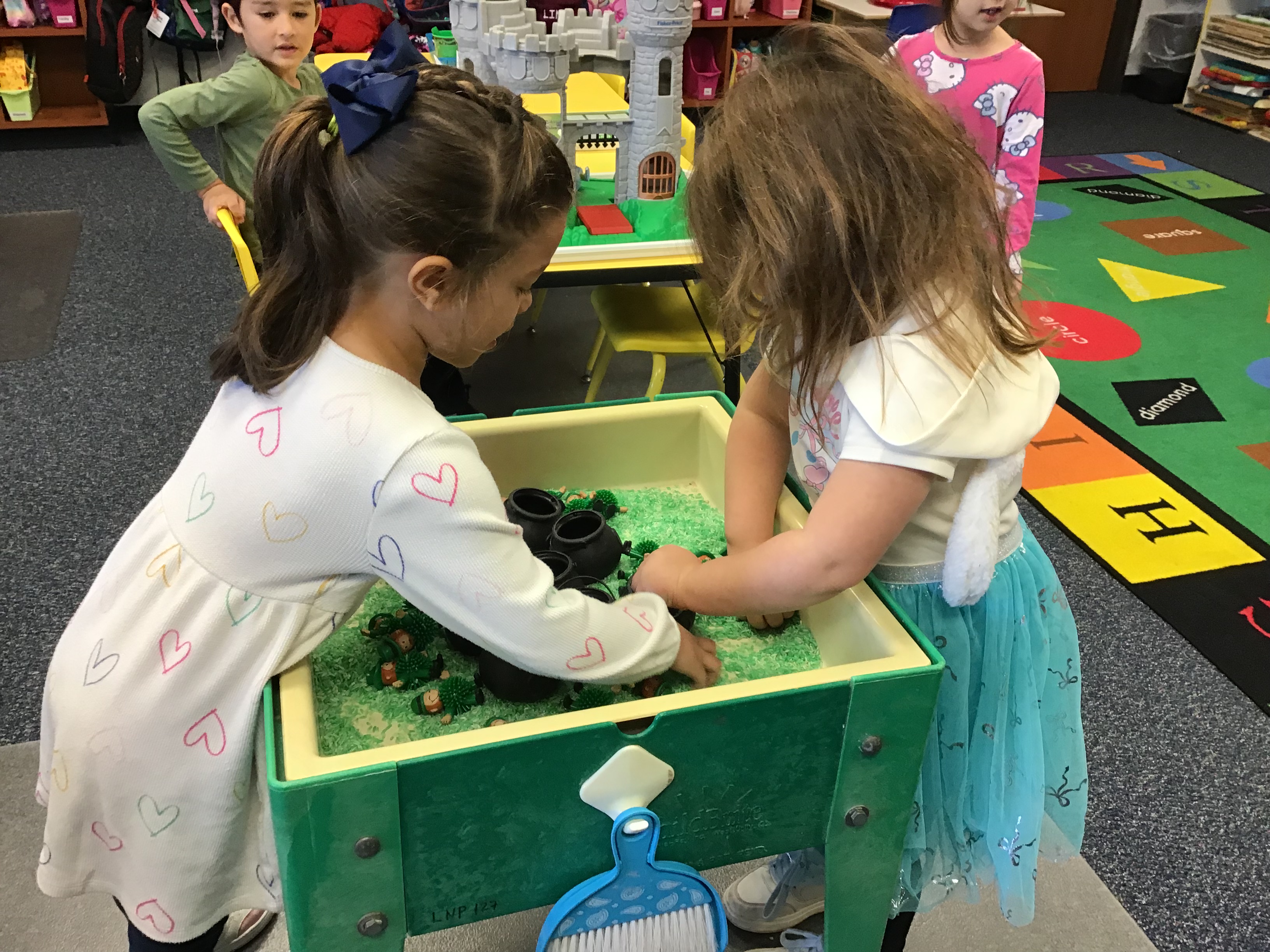 Two students playing at the sensory table filled with green rice , black cauldrons,  and leprechauns.