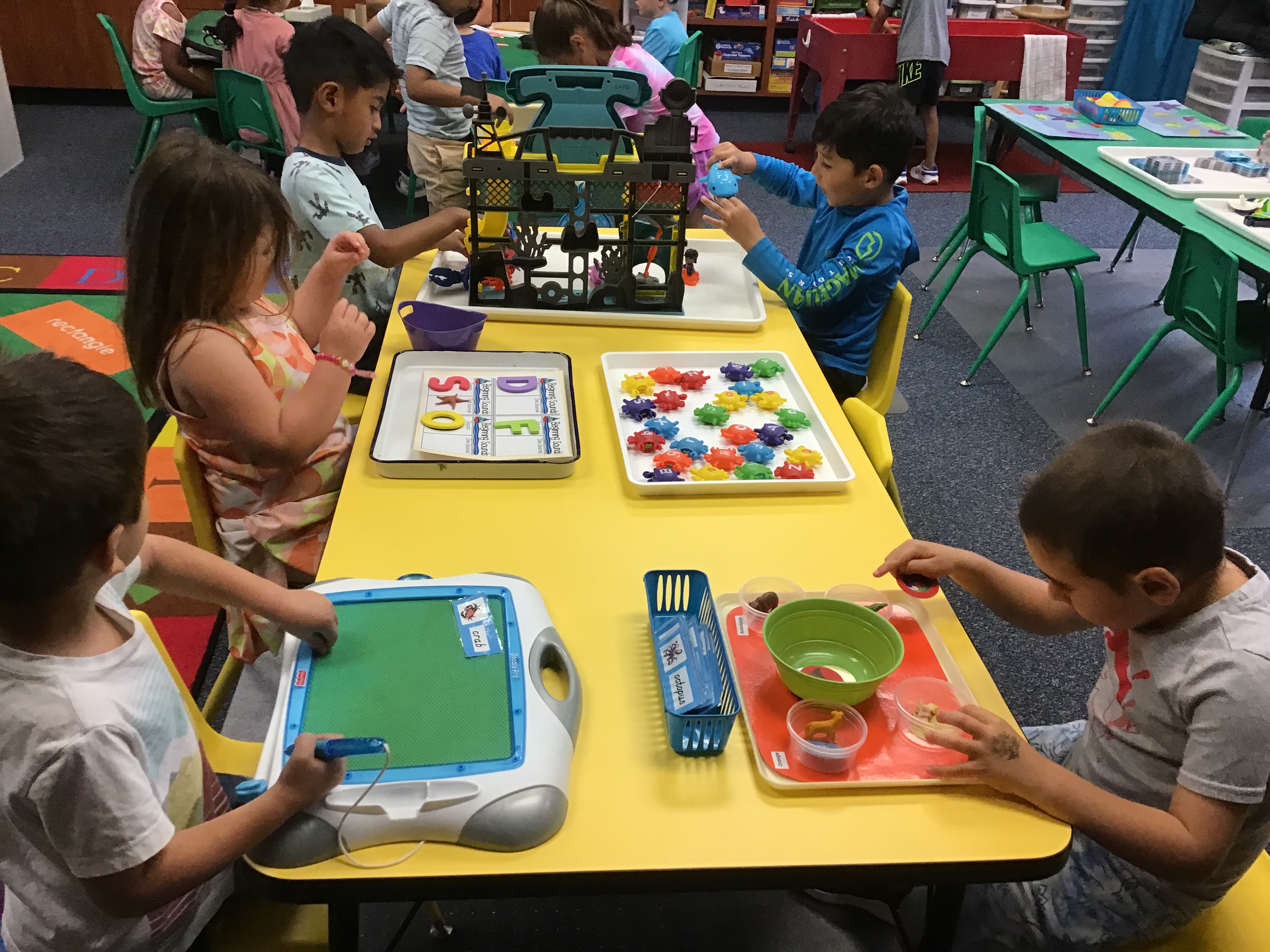 Two students at the phonics table writing theme words and sorting objects by first sounding letter.