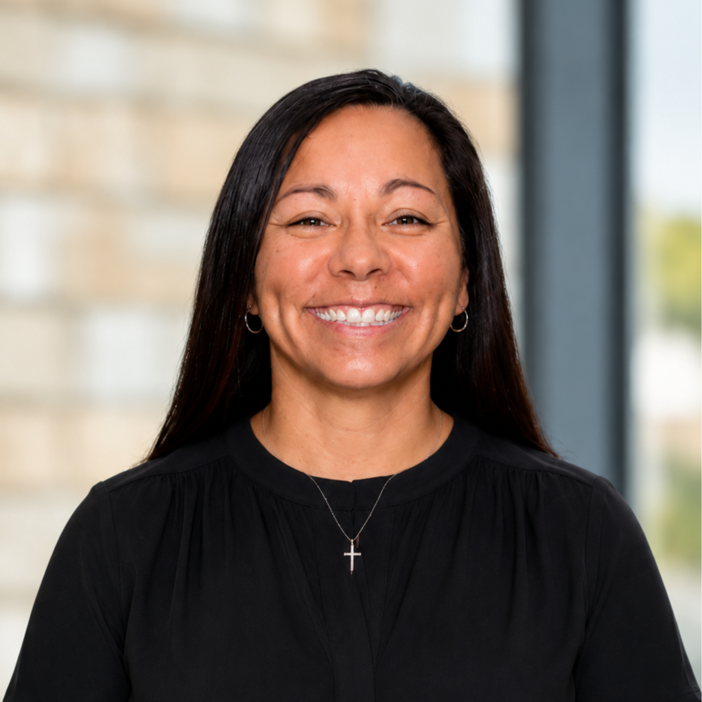 A woman with long dark hair smiles for a photo. She wears a black blouse and a cross necklace. Behind her, there is a blurry background.