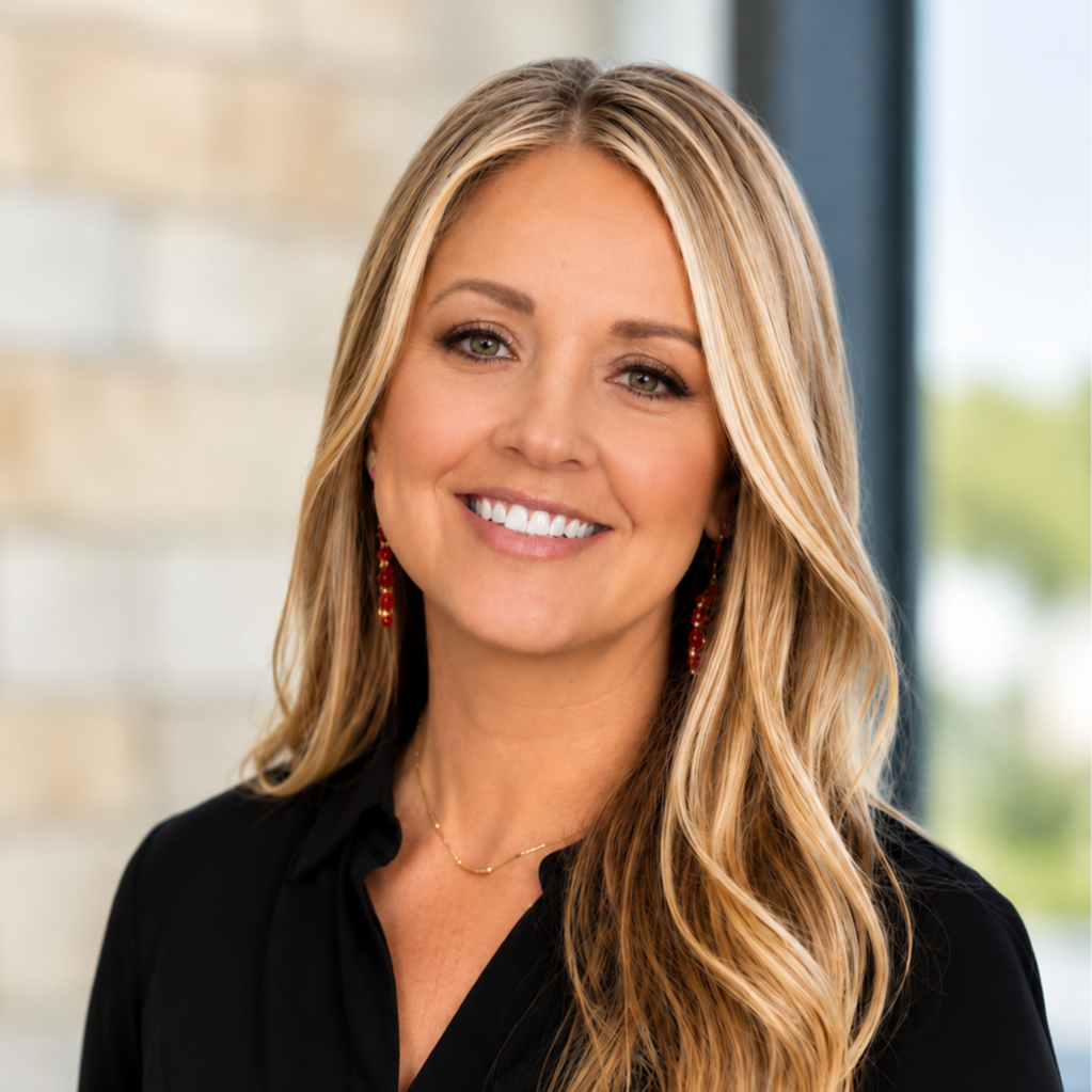 A woman with blonde hair smiles at the camera. She wears a black shirt and red earrings. Behind her is a wall.