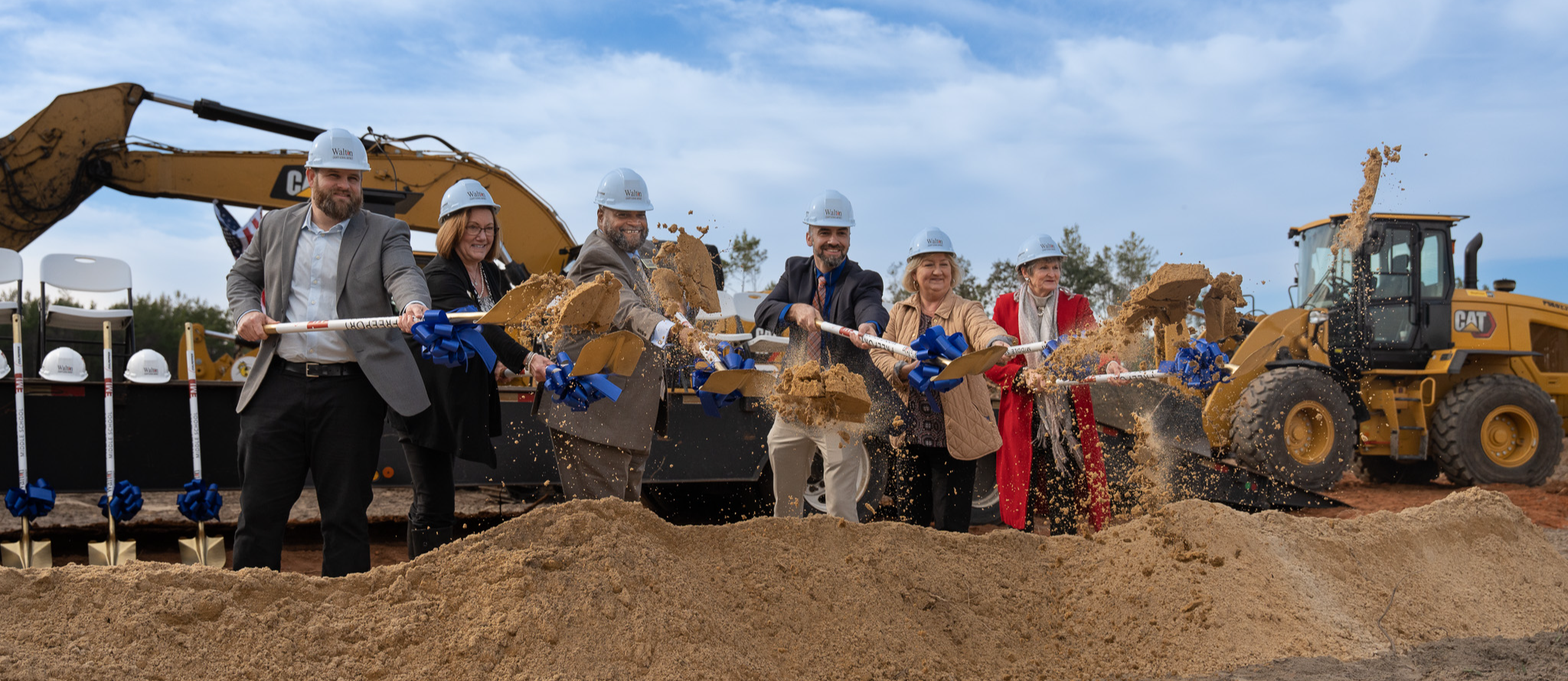 Superintendent Hughes and board members breaking ground on a new construction site, smiling in their hardhats as they lift their shovels