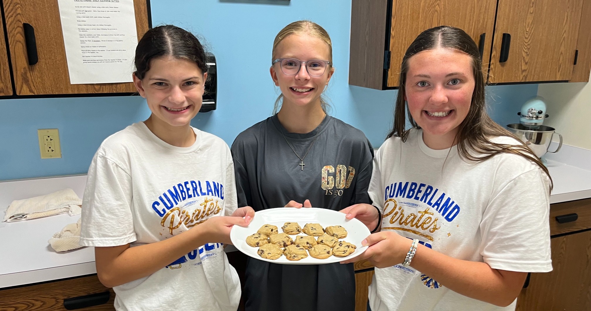 Three students are holding a plate of baked items. 