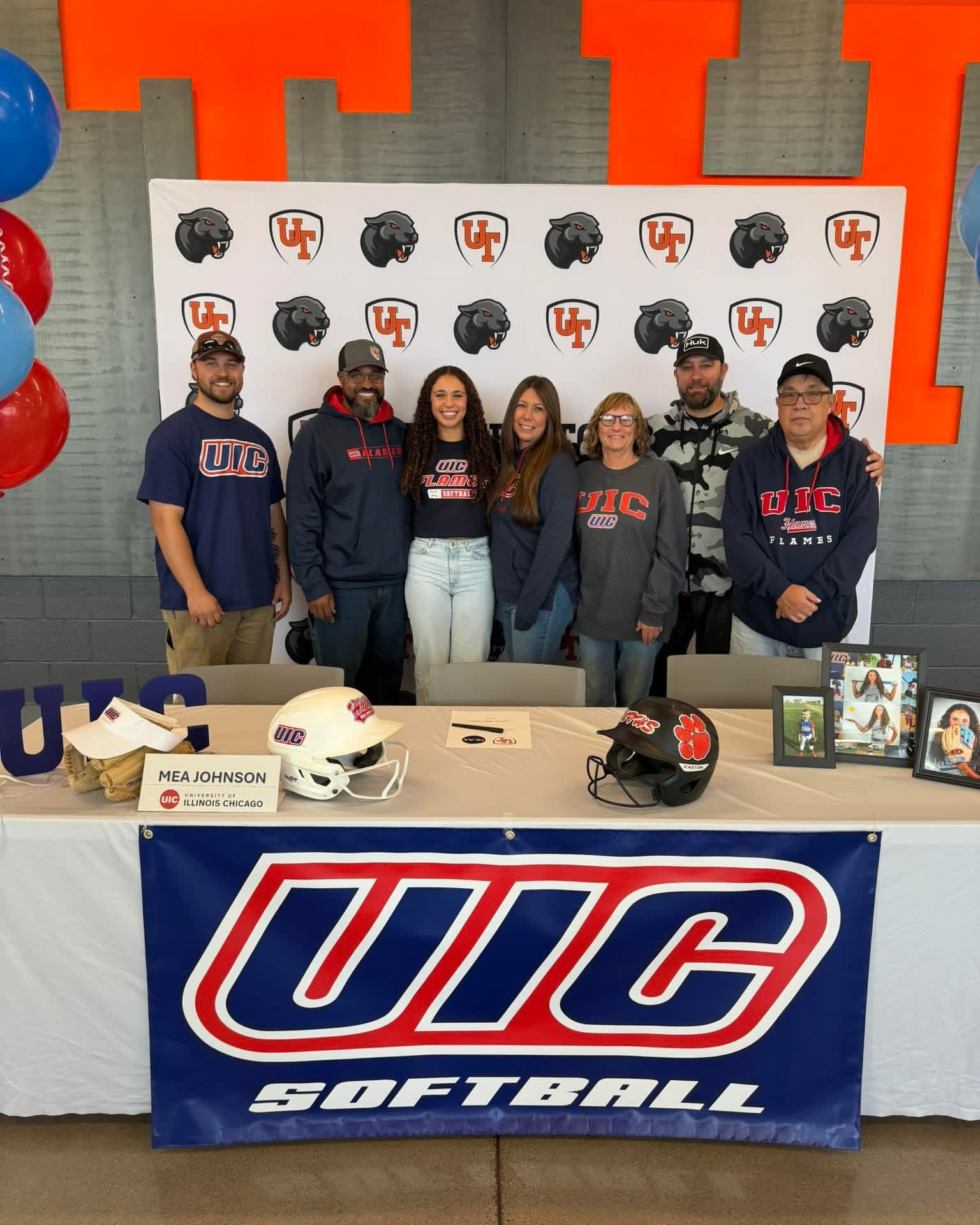 Image of table with softball swag from UIC and student with her family and on her NIL signing day. Red and blue balloons are visible as they are pictured in front of the panther backdrop.