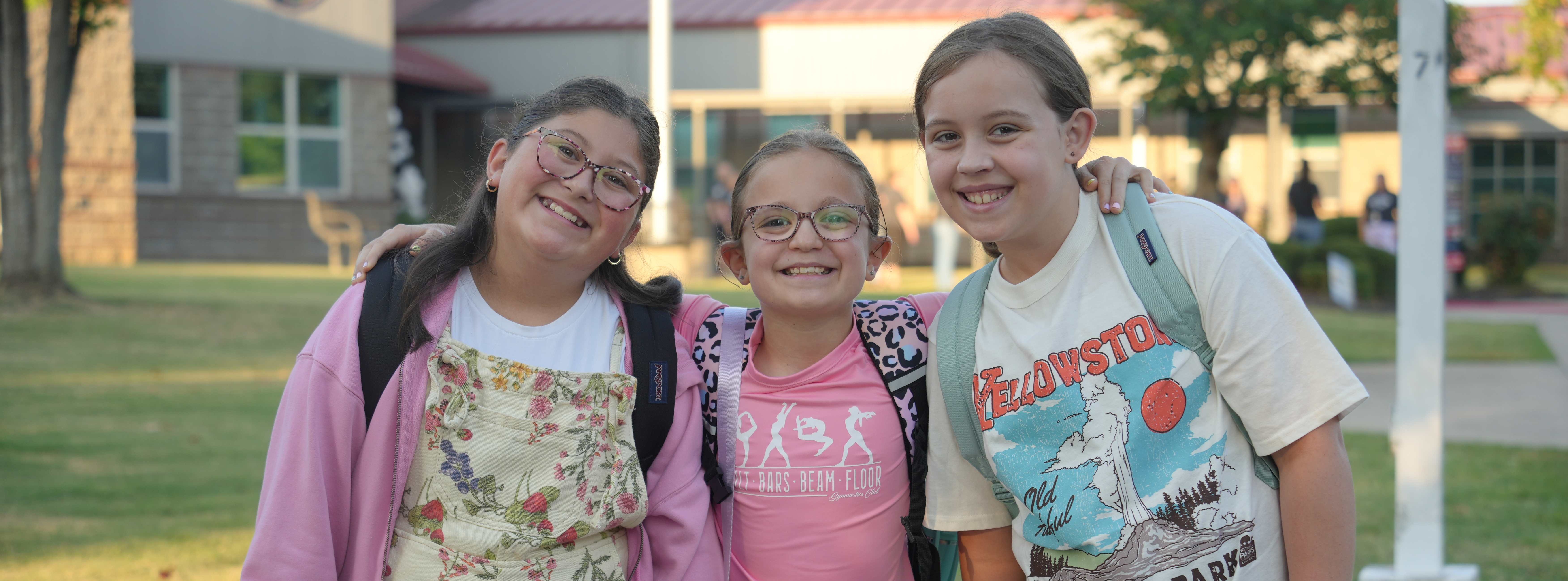 Three young people with backpacks in a schoolyard, standing together and smiling. The building behind them has red roofs.