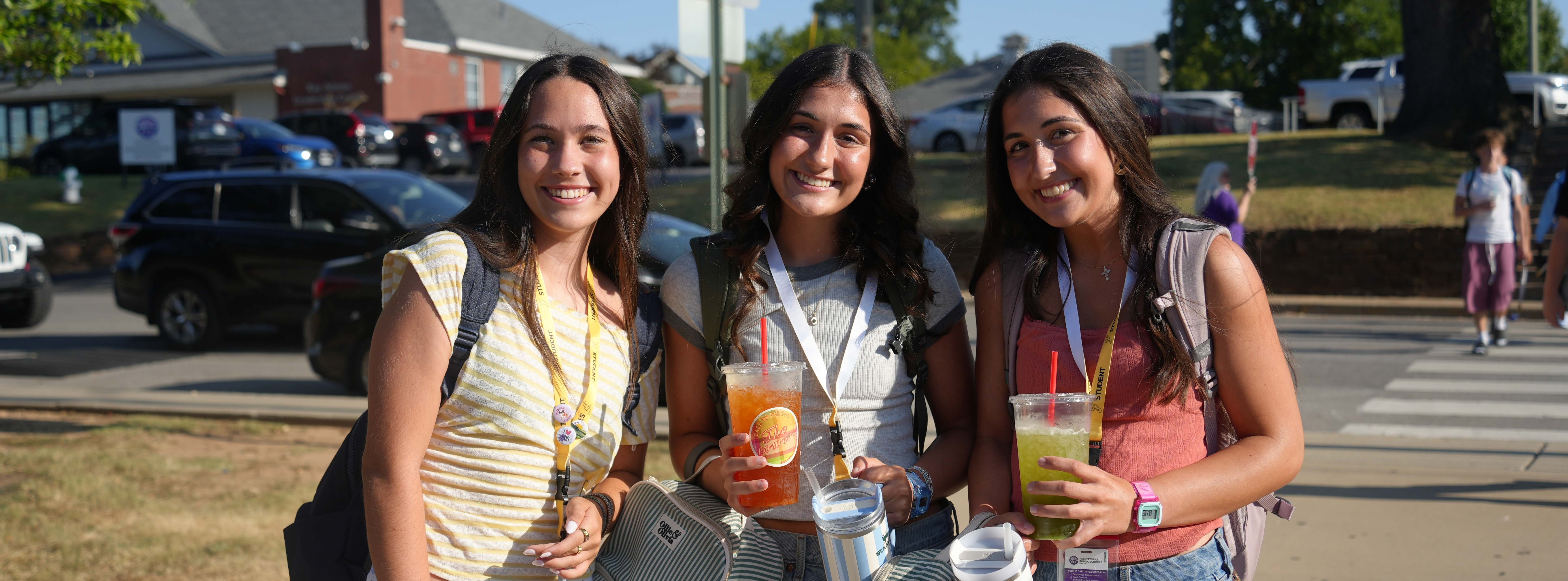 Three women standing on a sidewalk, each holding drinks, smiling at the camera. Behind them, cars parked, a grassy area, and a crosswalk.