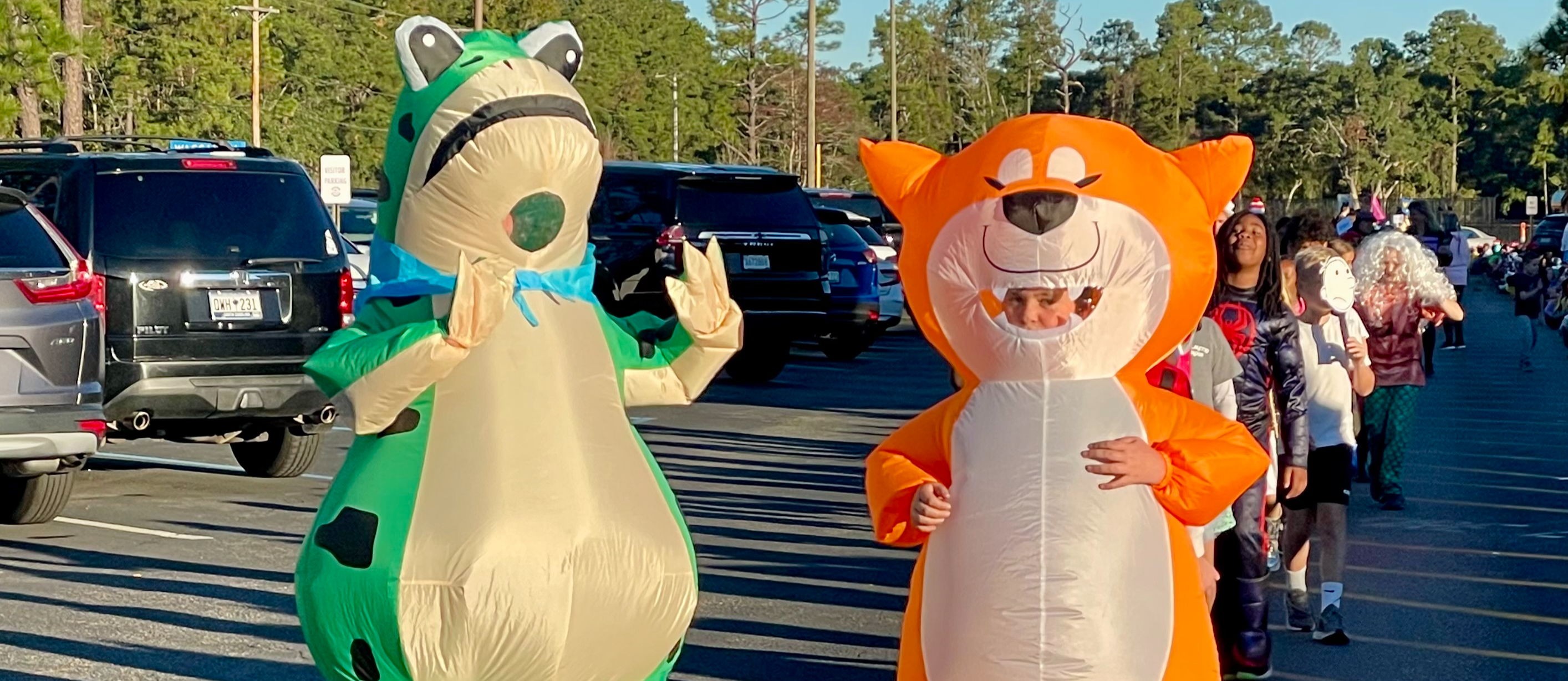 Two people walk outside in large inflatable costumes—one dressed as a green dinosaur and the other as an orange fox. A line of children in Halloween costumes walks behind them. The scene takes place in a parking lot under a bright blue sky.