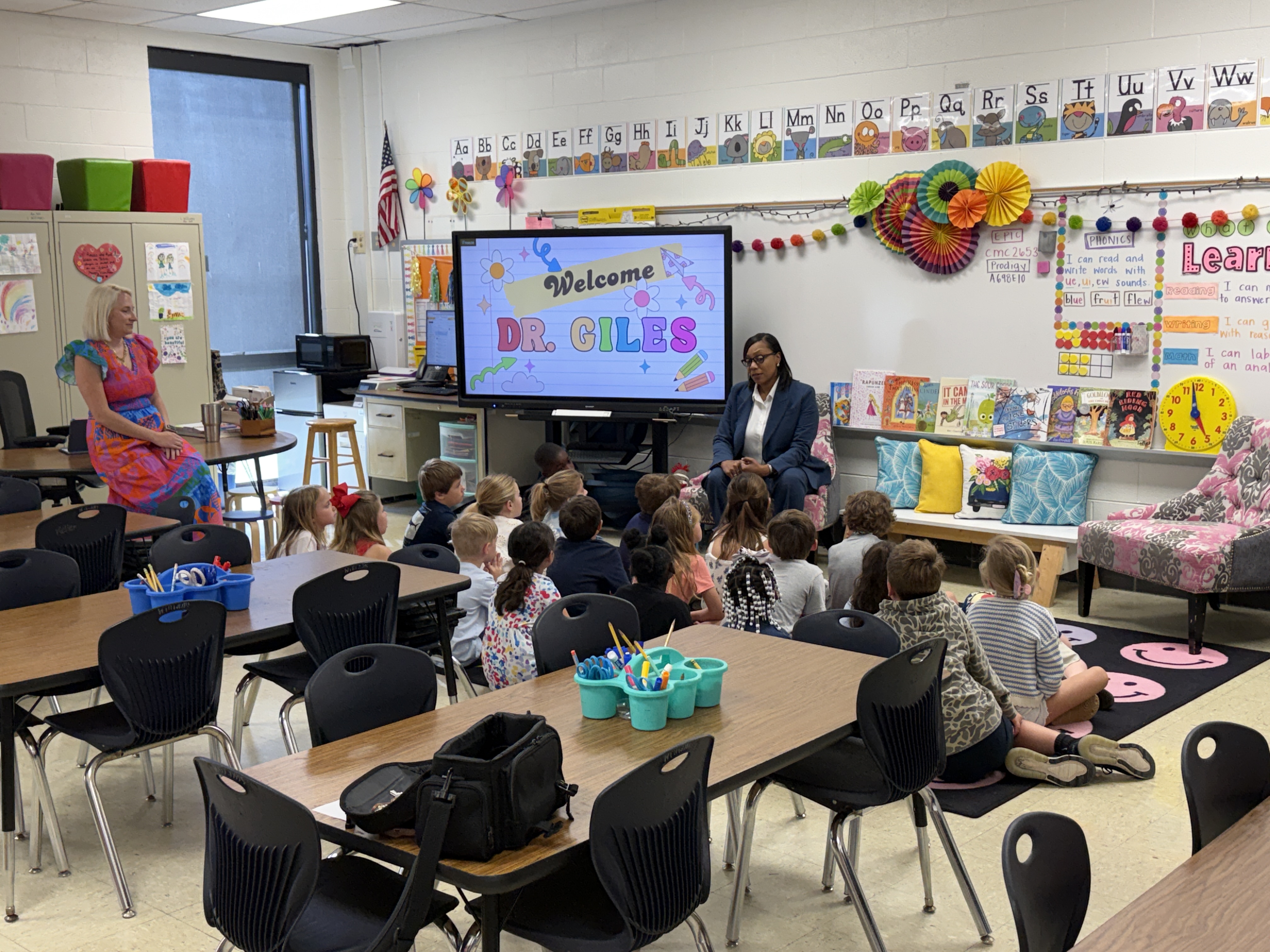 A guest reader sits at the front of the classroom reading a picture book while students sit attentively on the floor; a screen behind her welcomes a special guest.