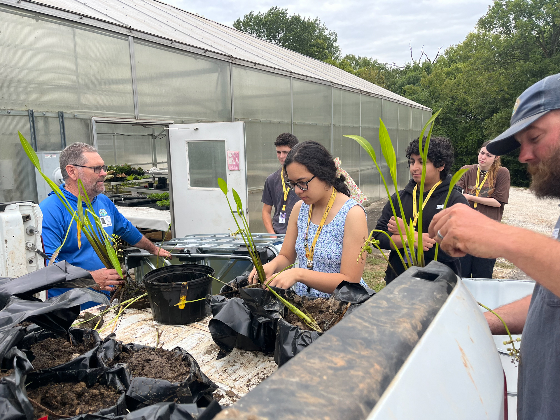 Several people examine potted plants outdoors. One holds a plant, another studies it, and two more look on.