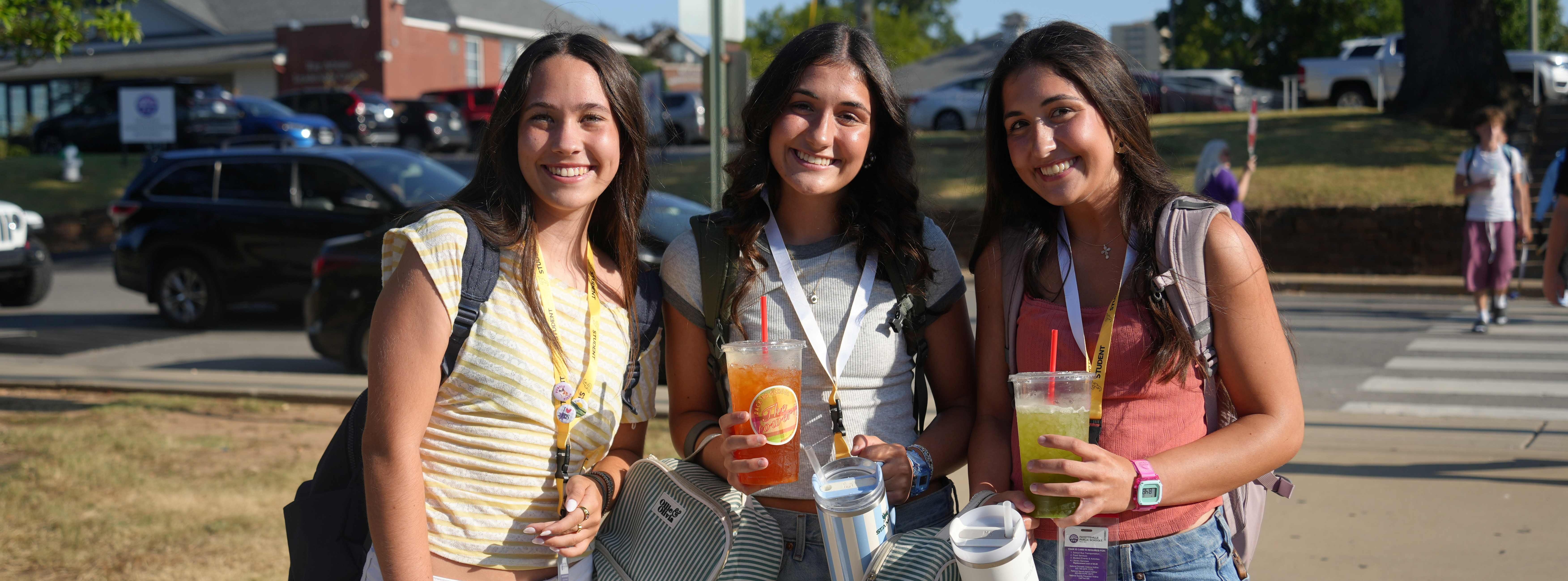 Three women standing on a sidewalk, two holding drinks. One wears a backpack and a bracelet, another carries a bottle.