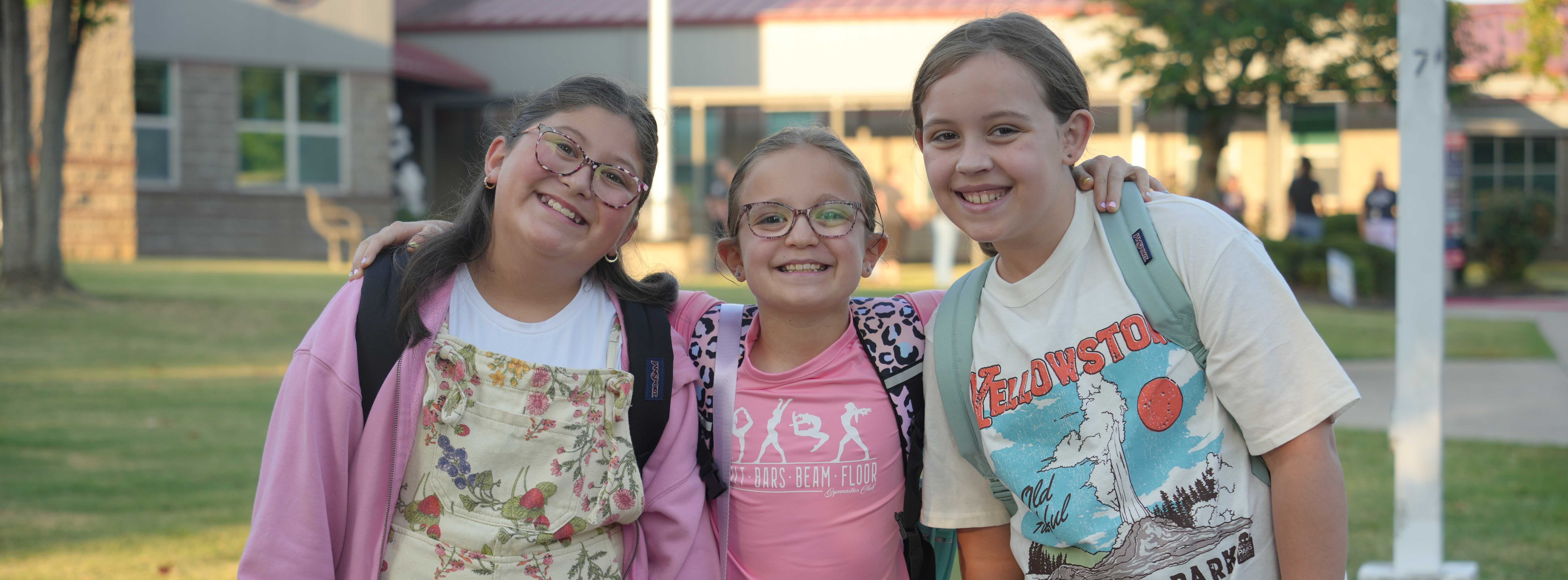 Three girls with backpacks in front of a building with a grassy lawn and trees.