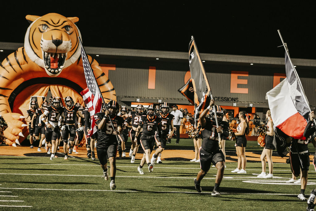 Football team marching on field with flags; large tiger graphic in background.