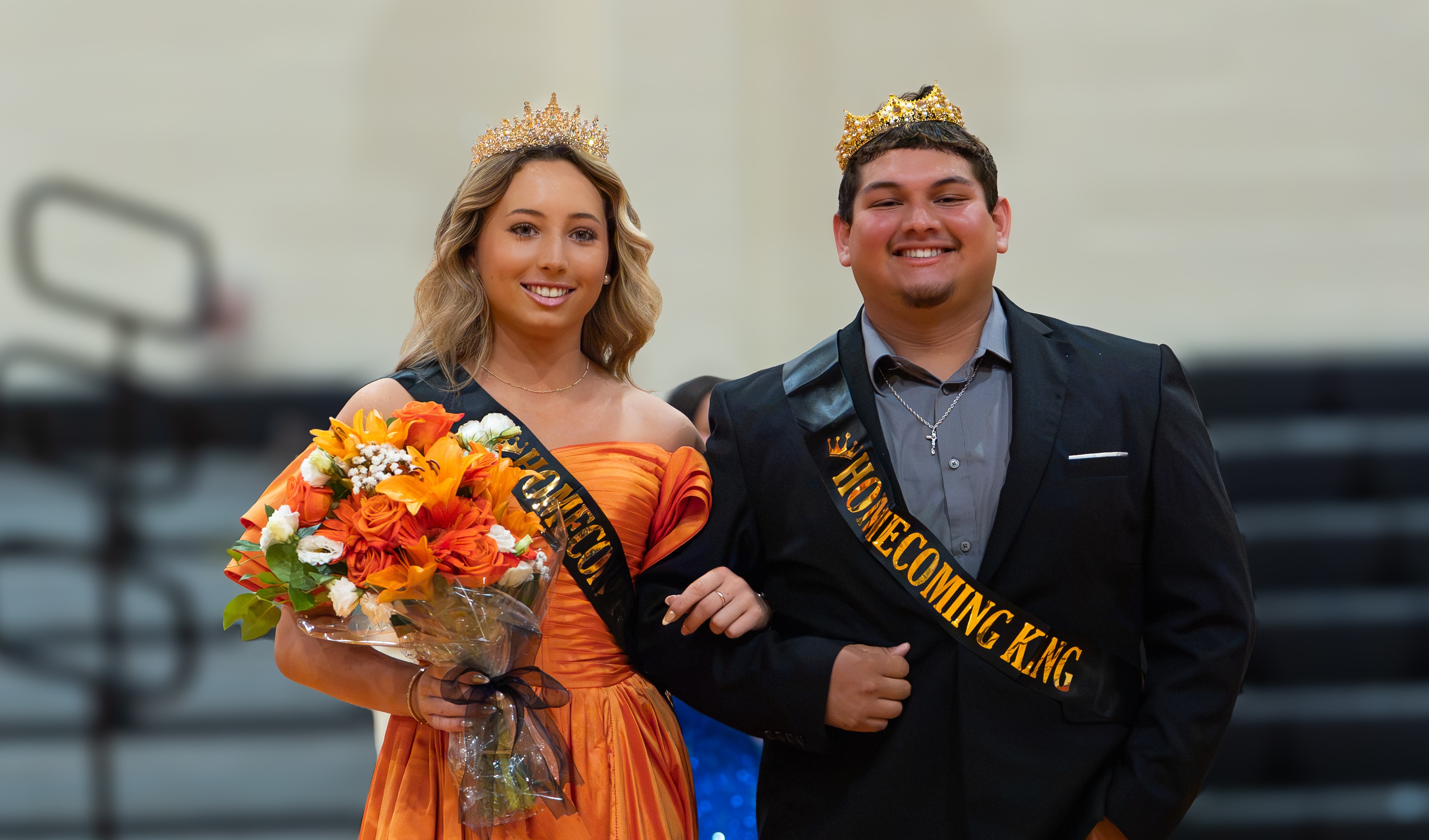 A woman in an orange gown and a man in a black suit stand on a basketball court.