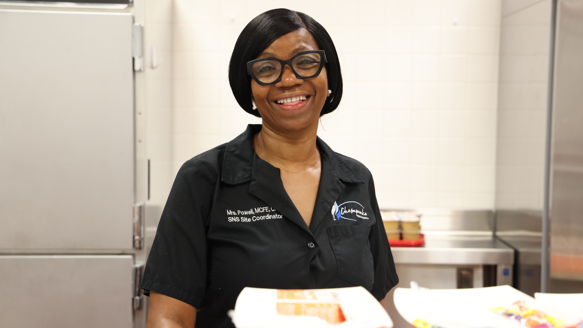 A school nutrition (lunchroom staff) worker smiles for the camera from behind the food service line.