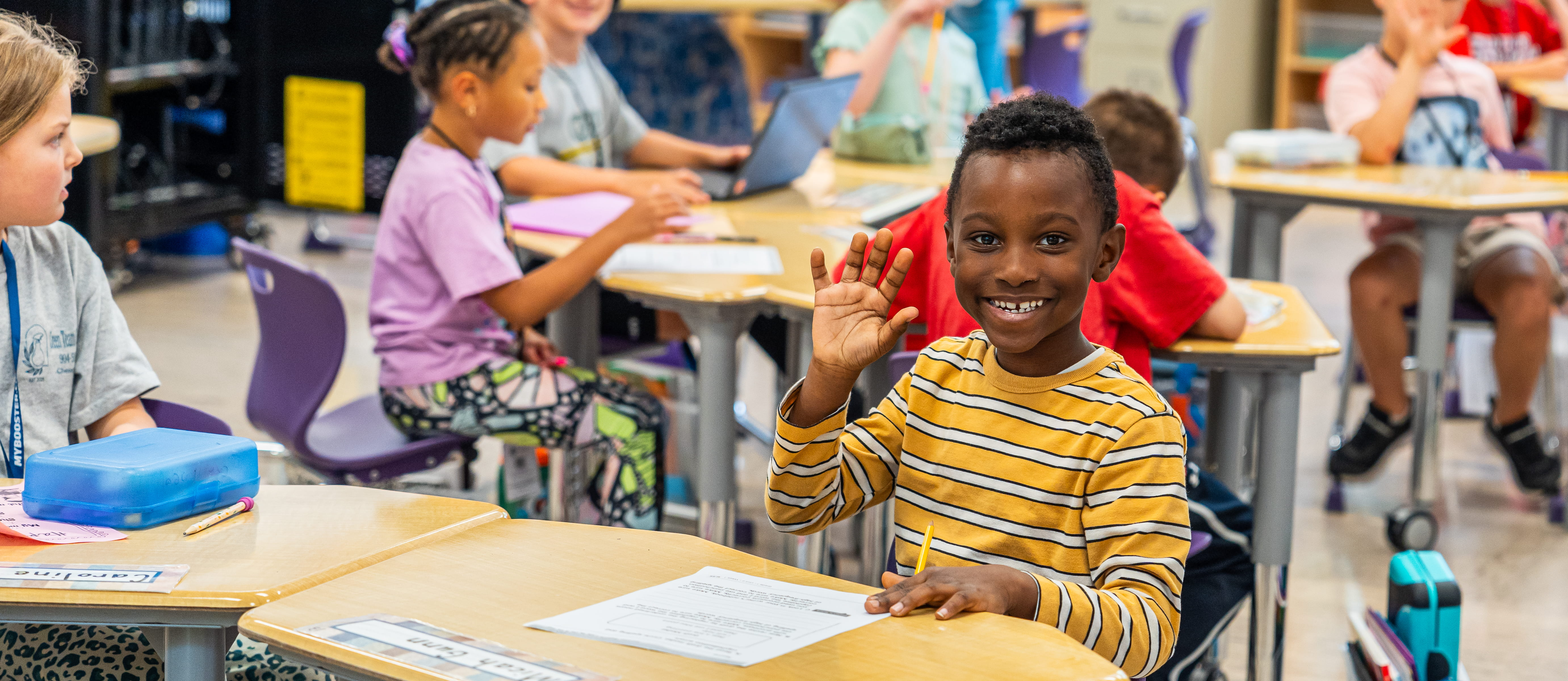 A student is an elementary classroom waves at the camera