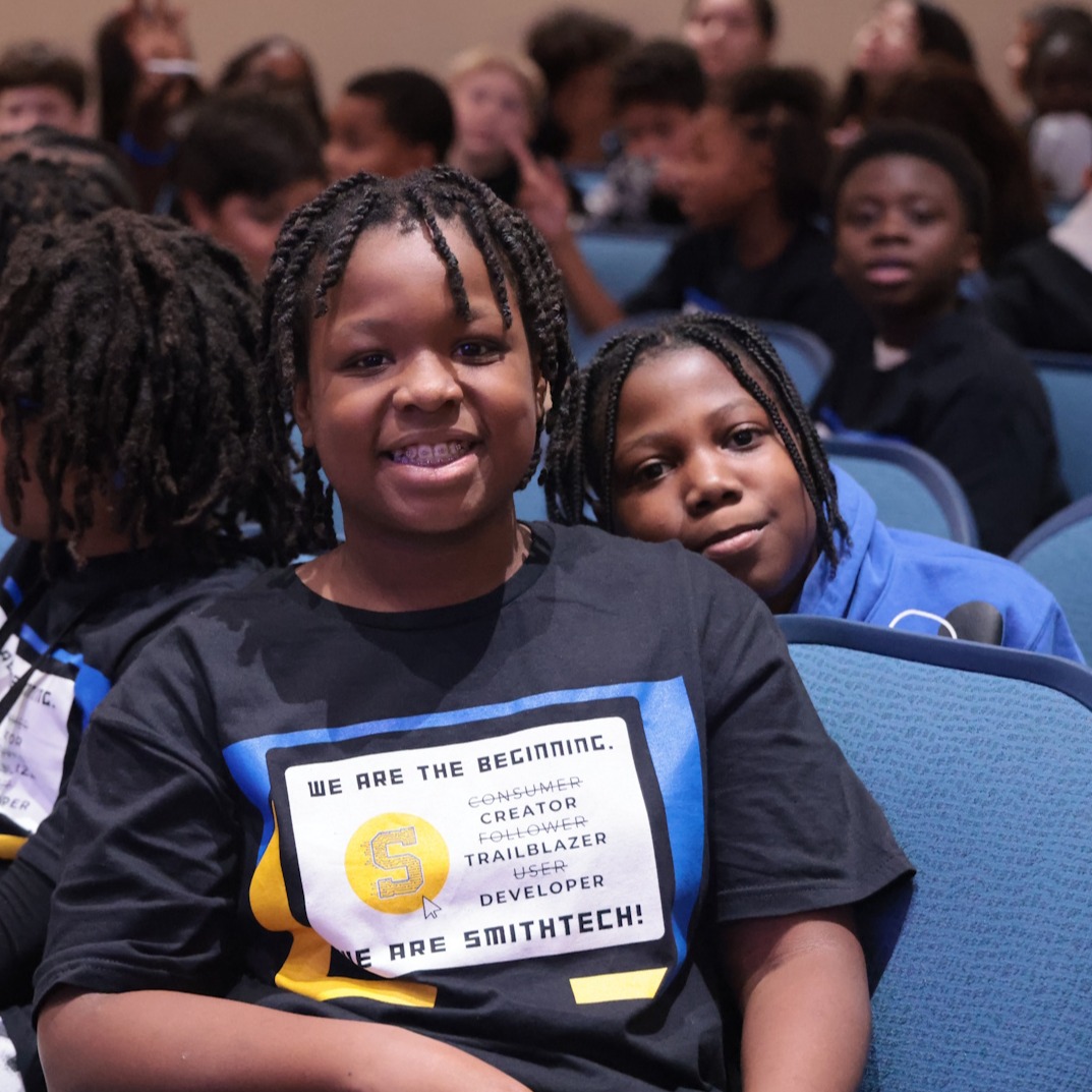 A SmithTech student wearing a SmithTech t-shirt smiles for the camera from her seat at the grand opening event for SmithTech.
