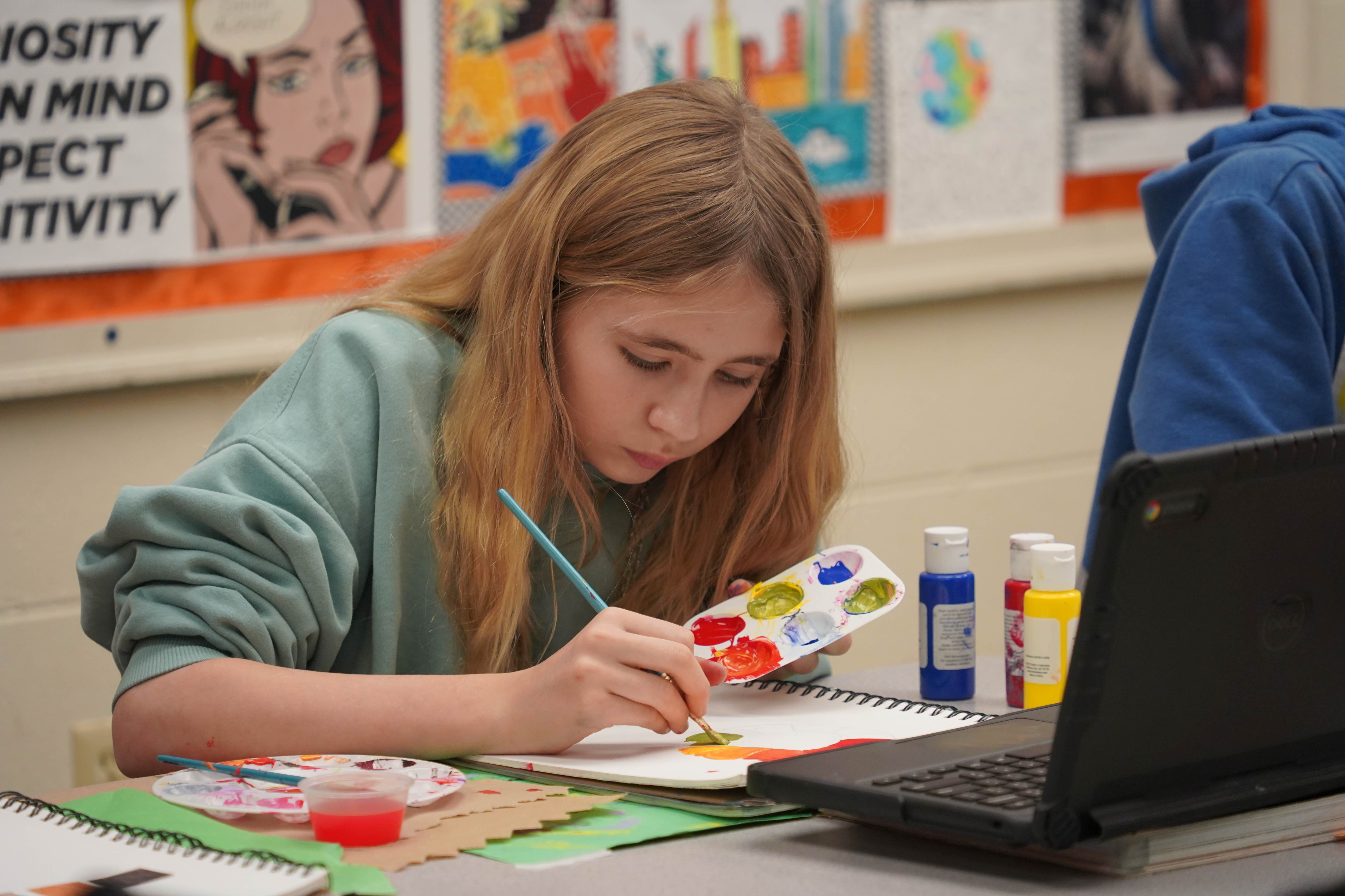 A student with long hair focuses on painting a palette. In front is a laptop. A wall displays motivational posters.
