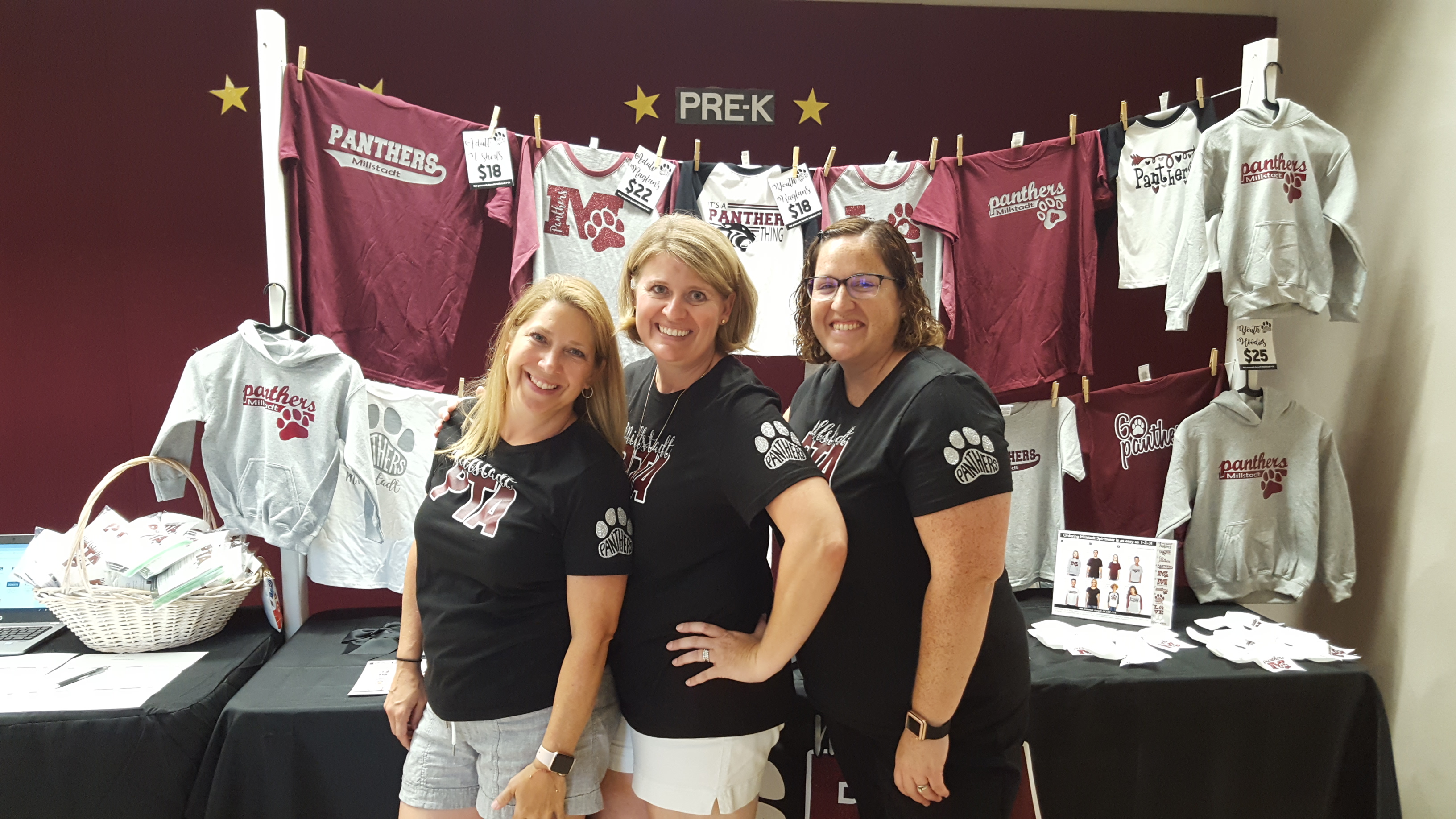 Three women, members of the PTA group, posing in front of their banner with their logo and a couple of shirts hanging at the back as part of their merchandise.