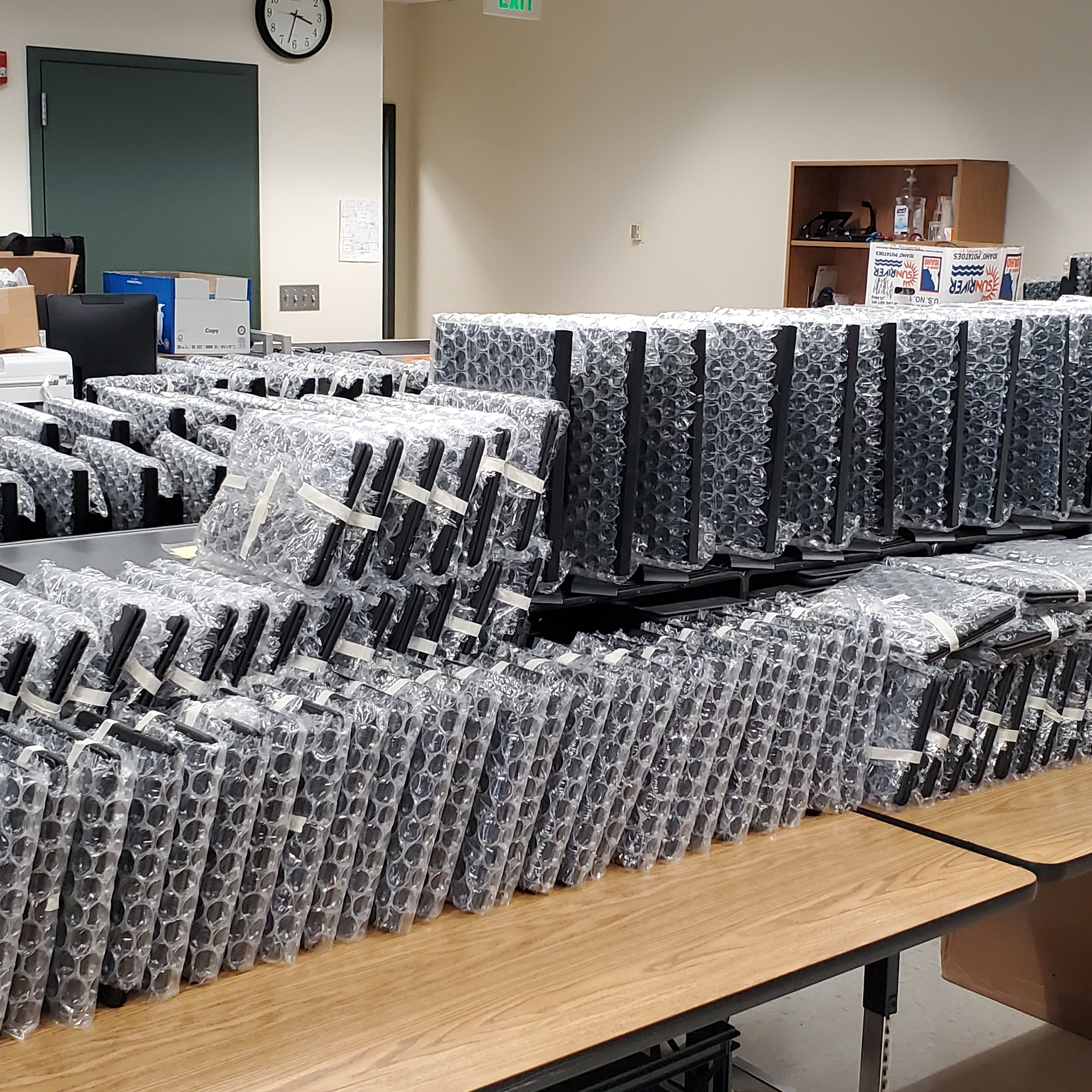 Roomful of Chromebooks or laptops as well as some monitors  wrapped in bubble wrap atop multiple tables. 
