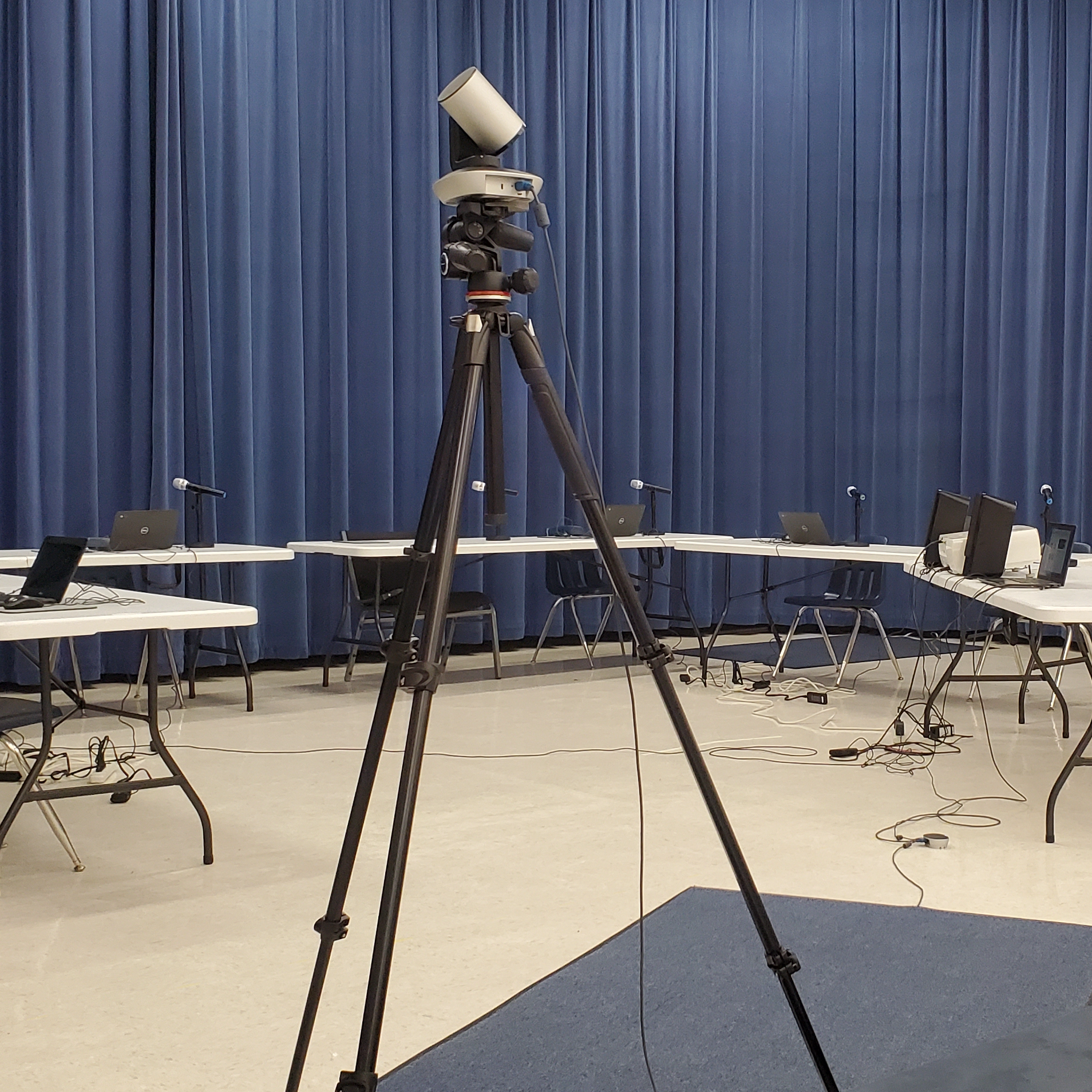 Setting up for school-board meeting at the middle school. The video camera is on a tripod in the center of the image with five viewable tables with laptops and microphones on them.