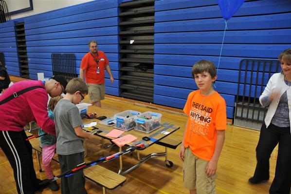STEM night: A boy looks at the camera. He has a shirt that says They states, They said I could be anything, so I became the boss.... An adult is behind him and another walking by. Three others are at the STEM station working on an activity.