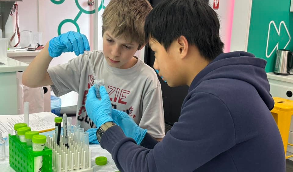 Two students in a science lab using a pipette to transfer a liquid into a test tube