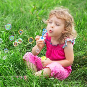 small child in a pink jumpsuit blows bubbles while sitting in the grass