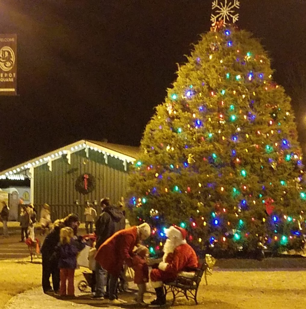 Santa Claus sitting on a bench talking to kids with a big lit Christmas tree and decorated Senior Center in background
