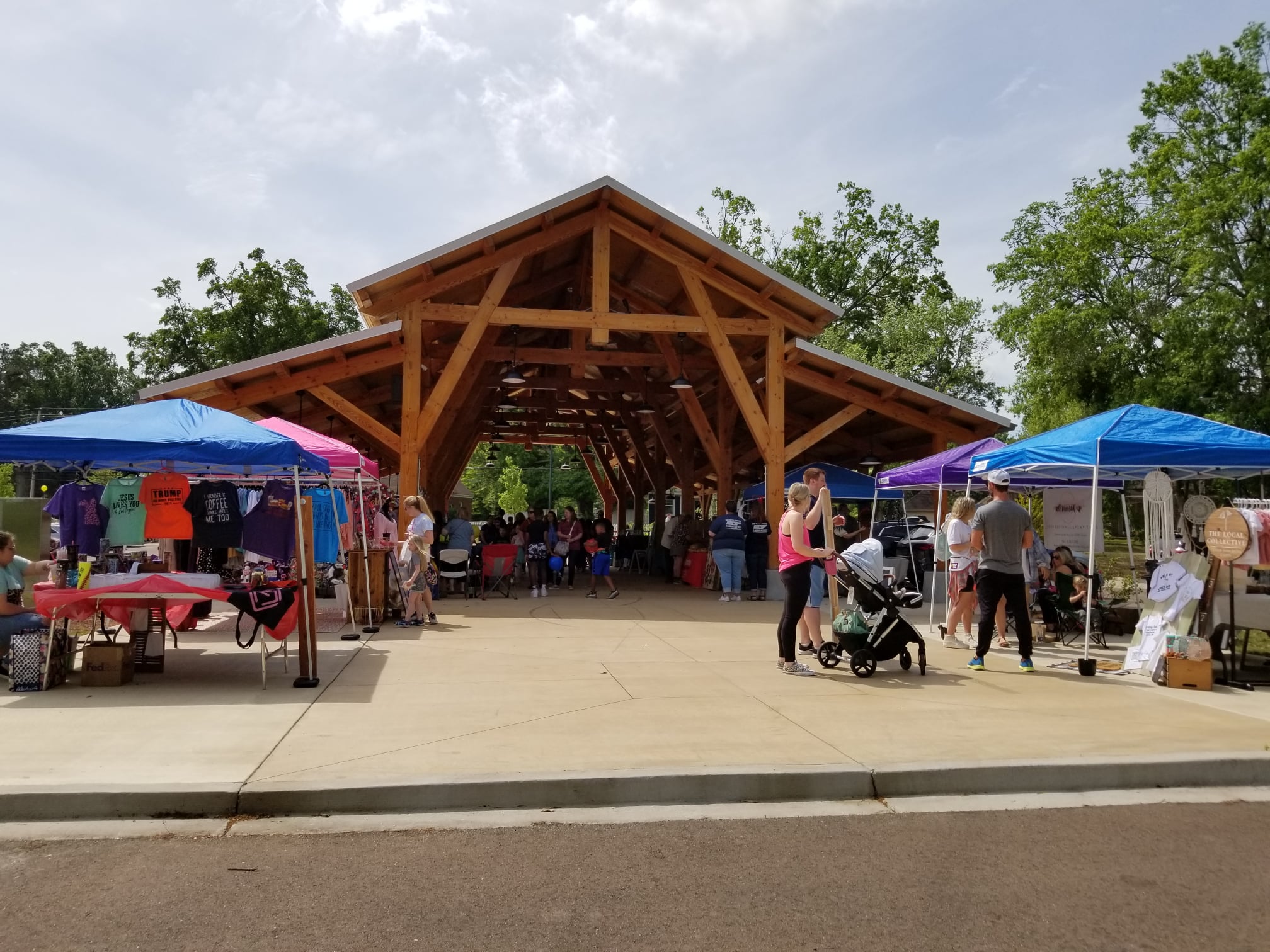 View of farmers market under wooden open-sided pavilion with blue tents on the sides with more vendors and people