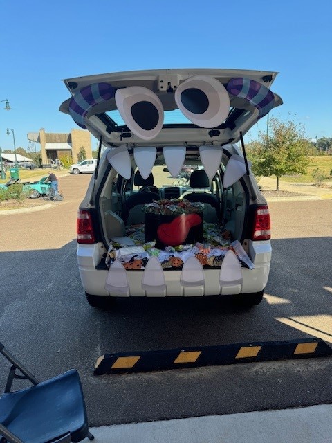 Decorated back of a white SUV with teeth, big googly eyes and a tongue bag with candy in the back. 