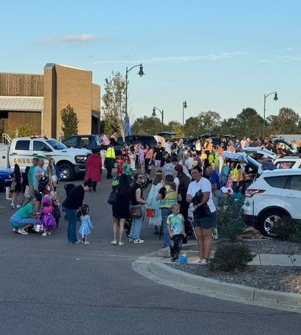 Groups of people dressed in costumes walking through trunk or treat event at park, with amphitheater in background