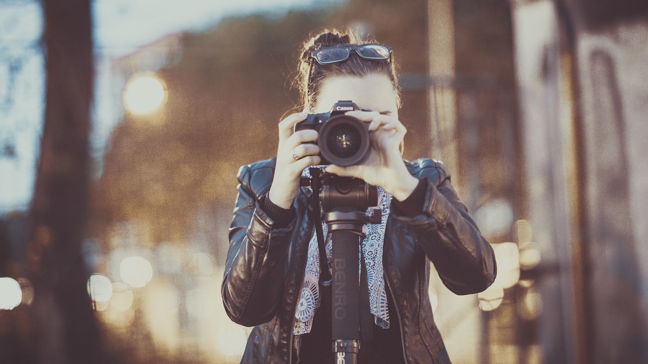 Picture of a lady in black leather jacket looking through a camera on tripod facing the camera