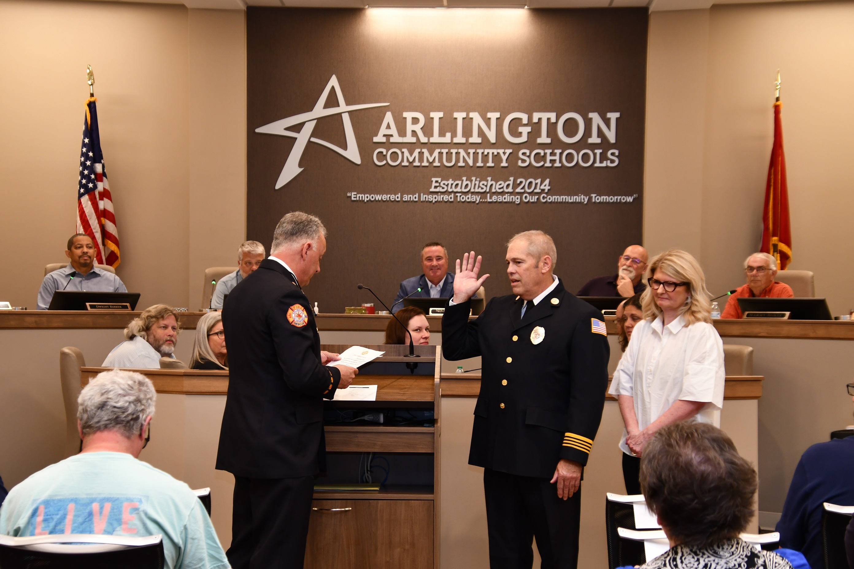 Picture of Fire chief in black dress uniform swearing in another fire chief in similar dress uniform with his wife beside him in a white blouse. All are in front of a raised dais with board members seated above and Arlington Community School lettering on back wall.